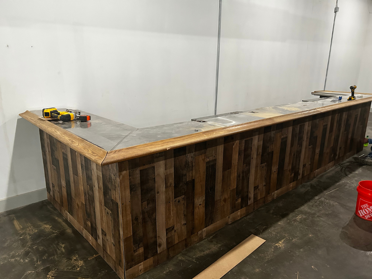 An L-shaped wooden bar with a metal countertop sits unfinished in a room with white walls and a concrete floor.