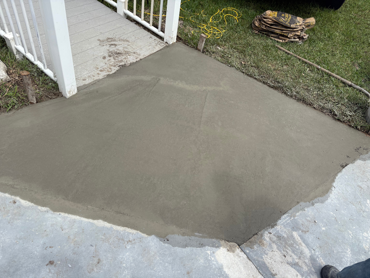A freshly poured concrete ramp transition connecting a wooden deck ramp to an existing sidewalk.