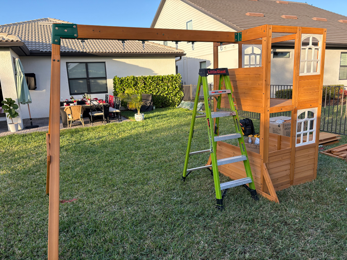 A partially assembled wooden playground structure with a green stepladder in a backyard.