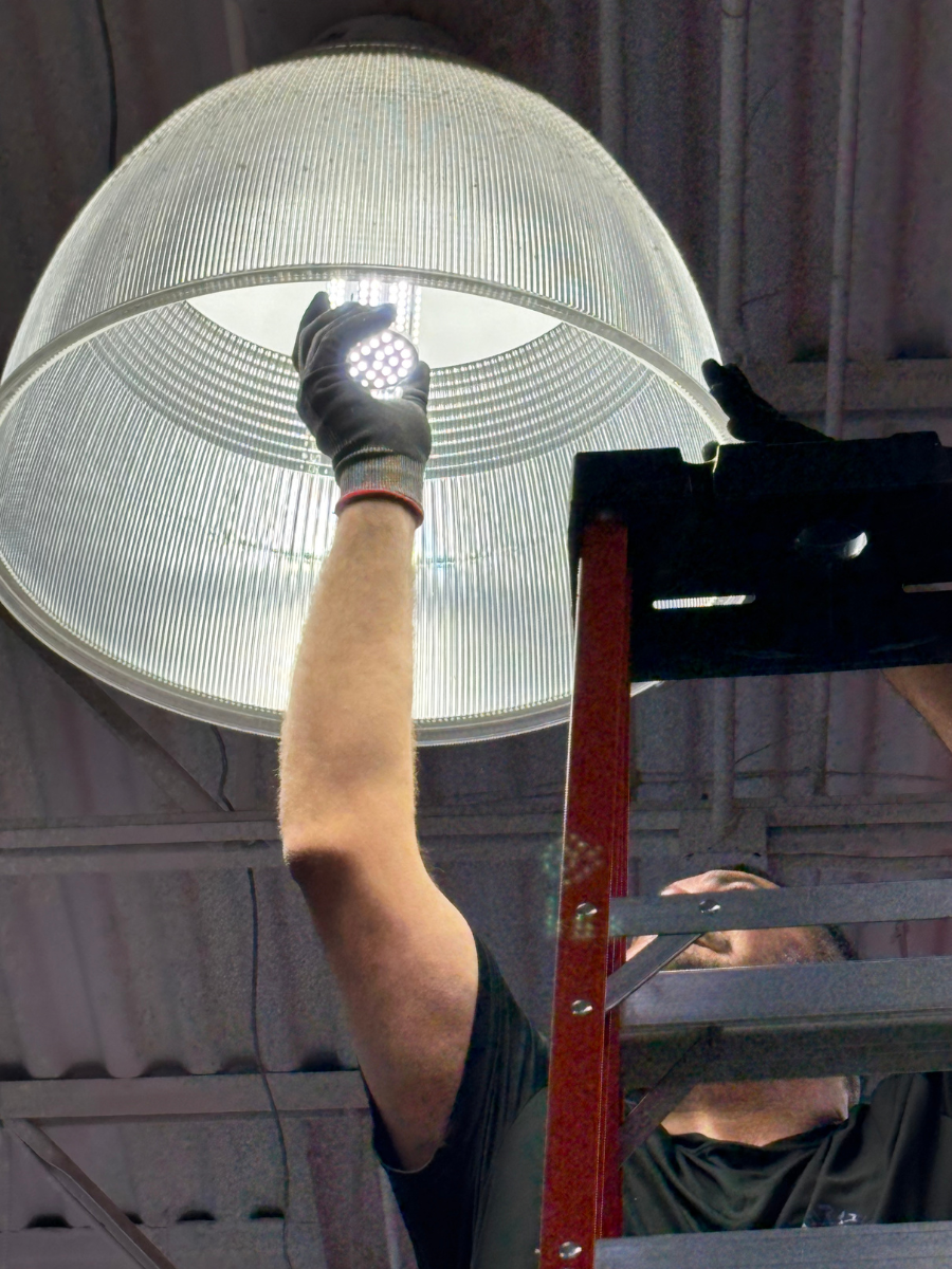 A person on a ladder wearing gloves changes a lightbulb inside a large, white dome-shaped pendant light.