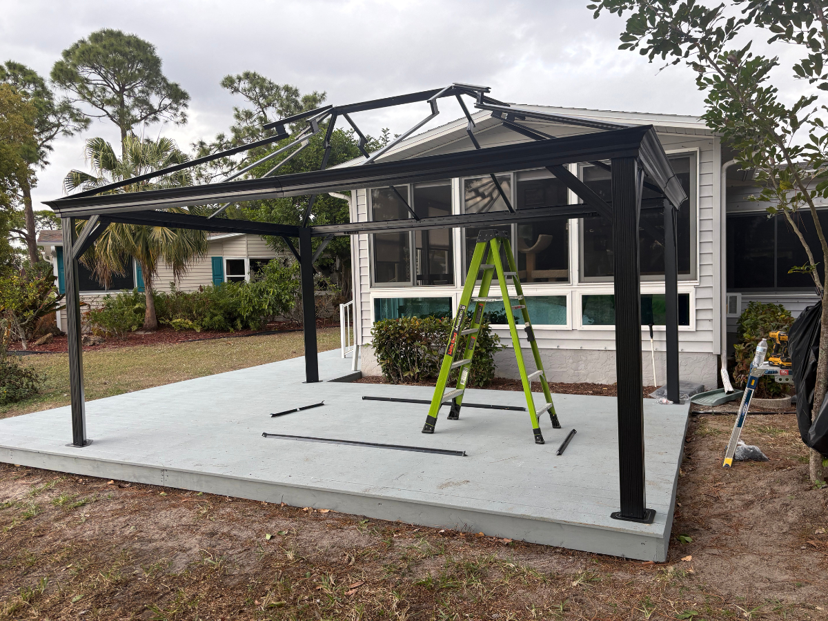 A black metal gazebo frame under construction on a gray concrete patio next to a house with a green ladder inside.