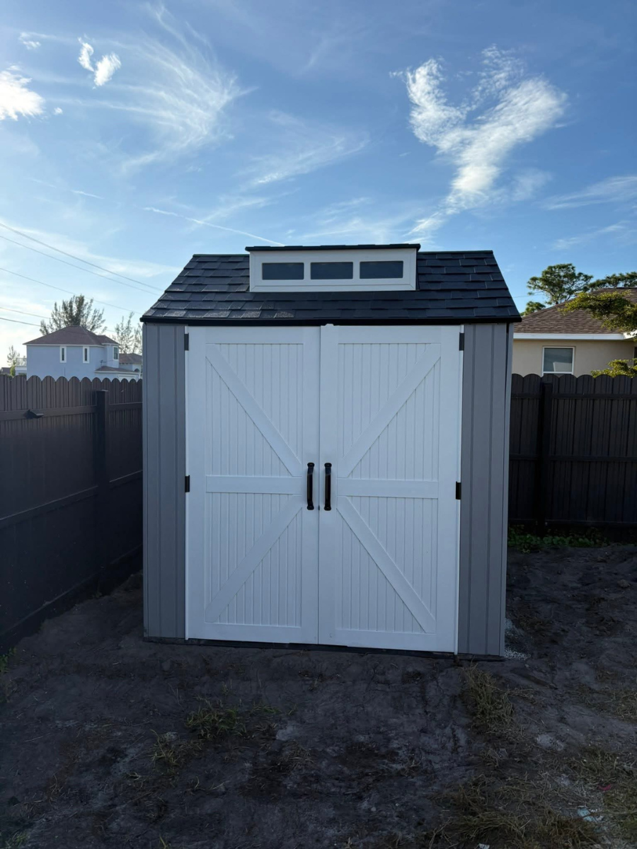 A grey storage shed with white double doors and a small roof window, set in an outdoor yard with fences in the background.