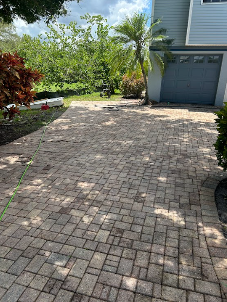 A paved stone driveway leads up to a gray house garage next to a palm tree and lush greenery under a cloudy sky.