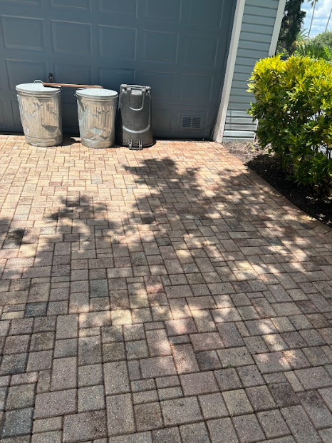 Three trash cans sit on a brown brick patio next to a gray building and a green shrub.