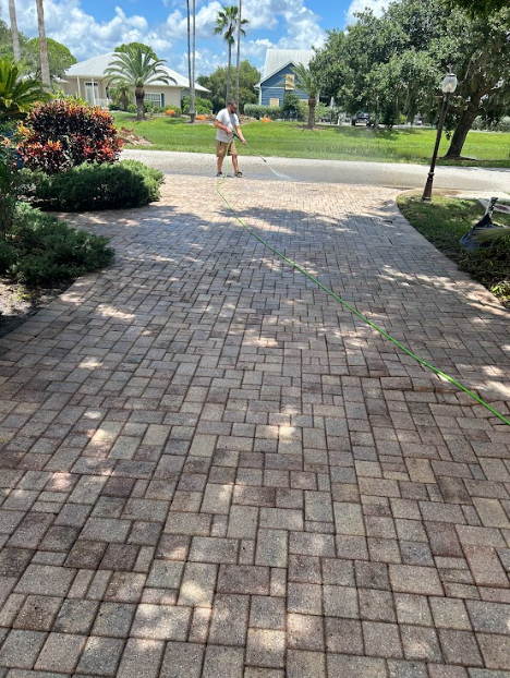 A person stands on a paved driveway, using a pressure washer to clean the interlocking brick surface.