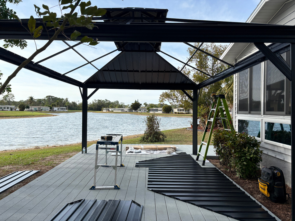 A partially assembled metal gazebo frame sits on a patio by a lake, with roof panels laid out for installation.
