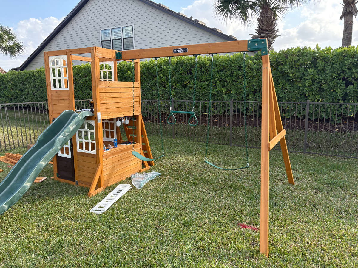 A wooden backyard swing set with a slide and a playhouse structure, positioned on a grassy lawn.