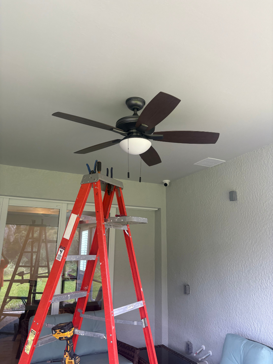 A ceiling fan installed in a room, with a red ladder and a power drill placed beneath it.