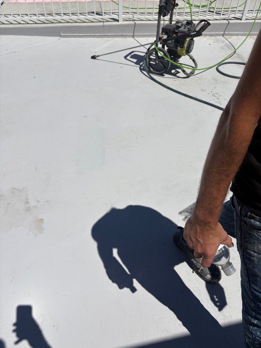 A person stands on a light gray surface holding a clear bottle, with a pressure washer nearby and shadows on the ground.