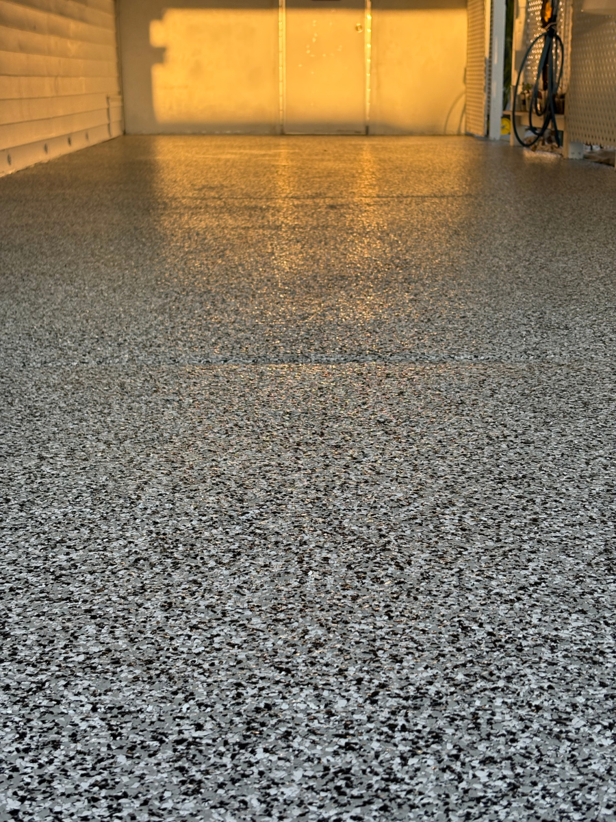 A garage floor with a smooth, speckled grey, white, and black epoxy finish, reflecting warm light from the back wall.