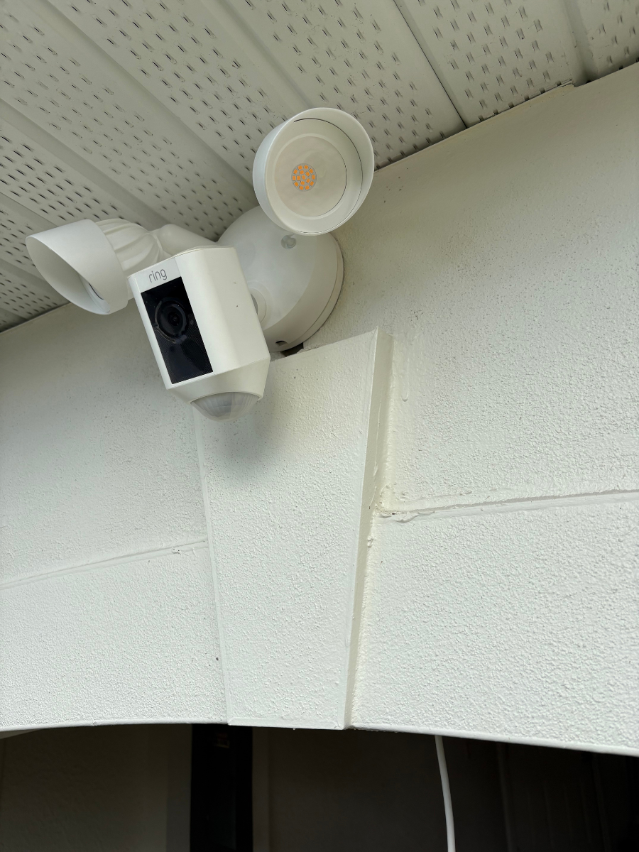 A white Ring Floodlight Cam mounted on a light-colored, textured stucco wall under an exterior soffit.