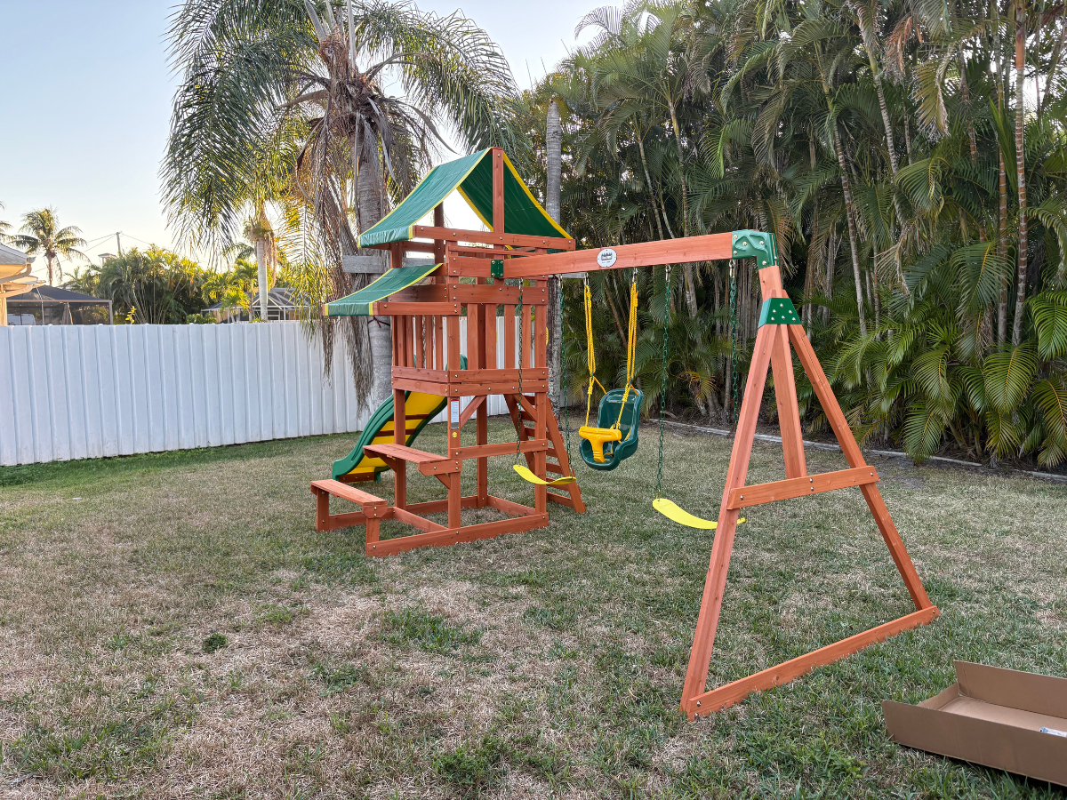A wooden playset with a green roof, slide, and two swings in a grassy backyard near a white fence.