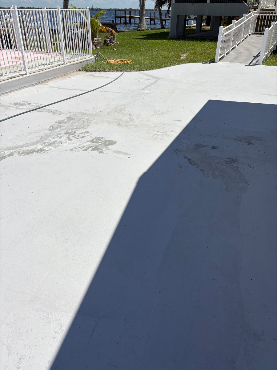 Concrete patio with a large shadow covering half the surface, looking toward a grassy yard, fence, and waterfront.