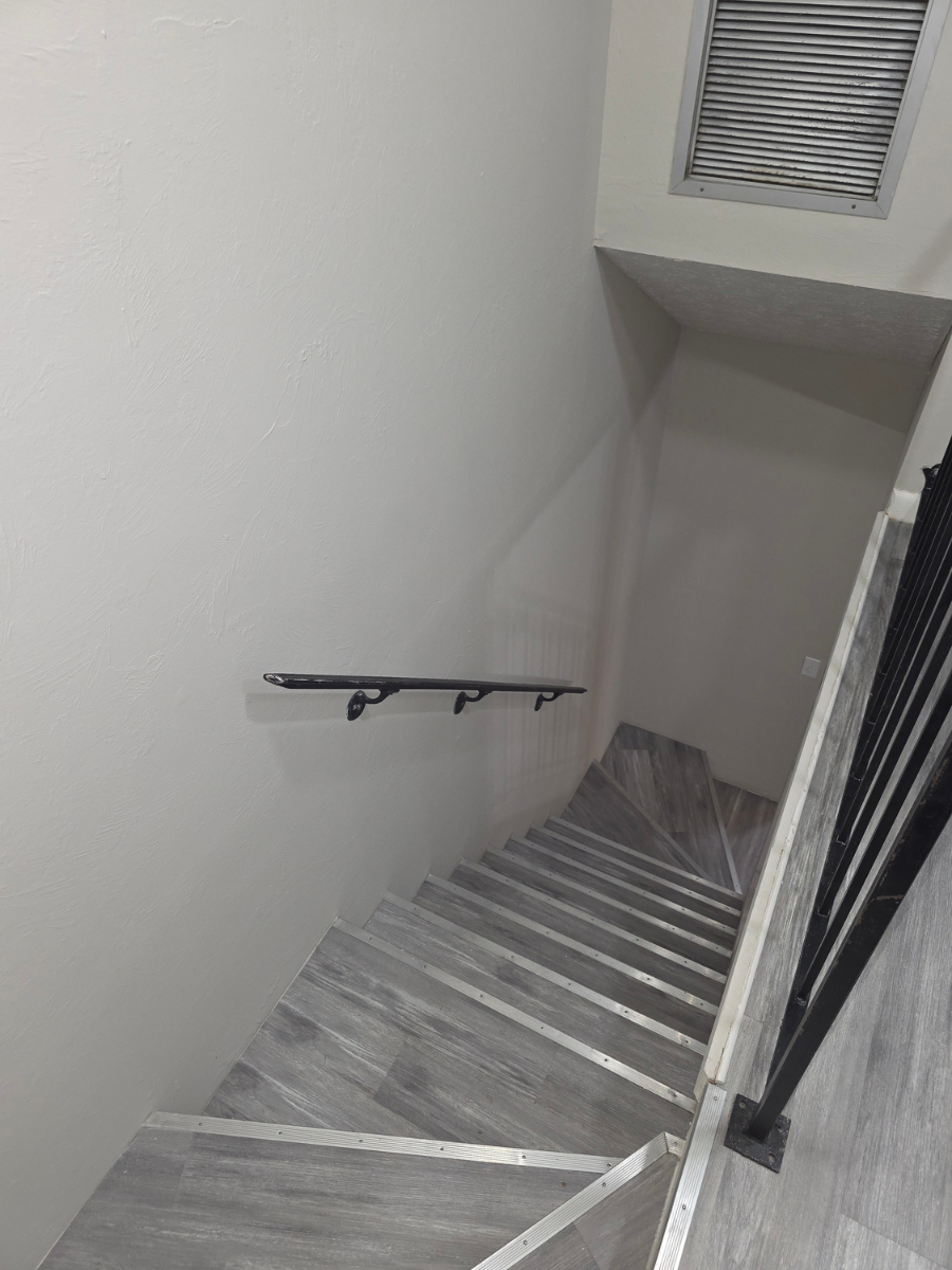 Downward view of light-colored wooden stairs with a black metal handrail and railing.