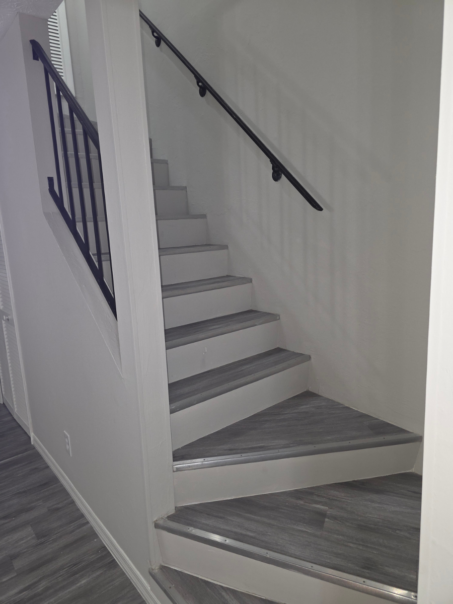 Indoor staircase with grey wood-look treads, white risers, black metal railings, and a black handrail on a white wall.