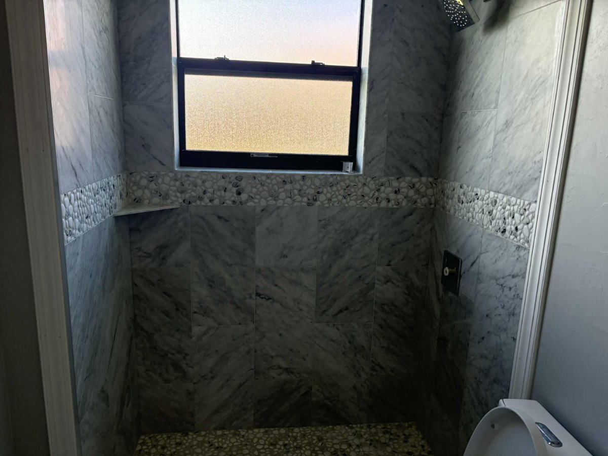 A small shower stall with gray marbled tile walls, a decorative pebble tile border, a window, and a white toilet nearby.