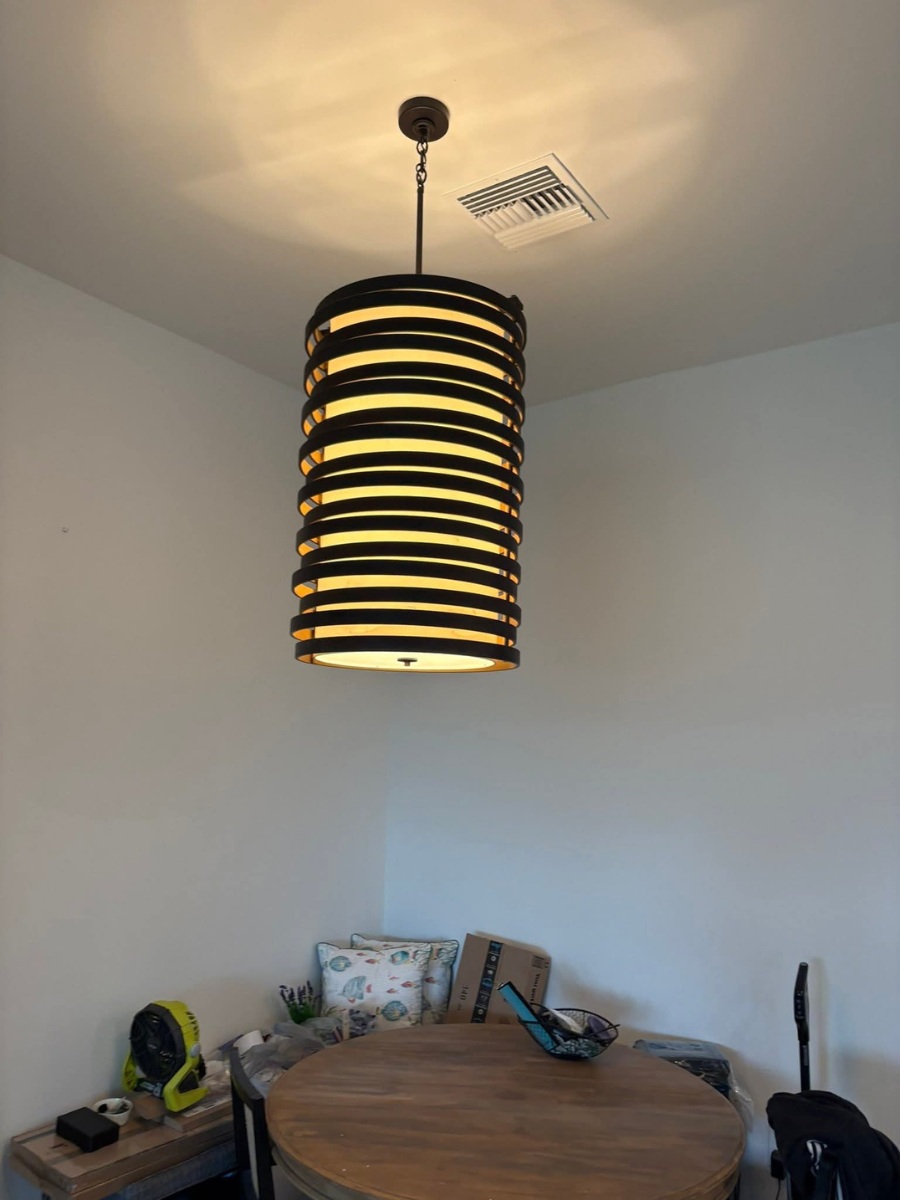 A modern, cylindrical pendant light with horizontal dark slats hangs above a wooden dining table in a neutral-toned room.