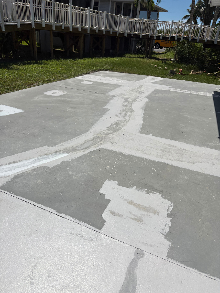 Concrete patio surface with light-colored patches and filled cracks, set in a yard with a wooden deck in the background.