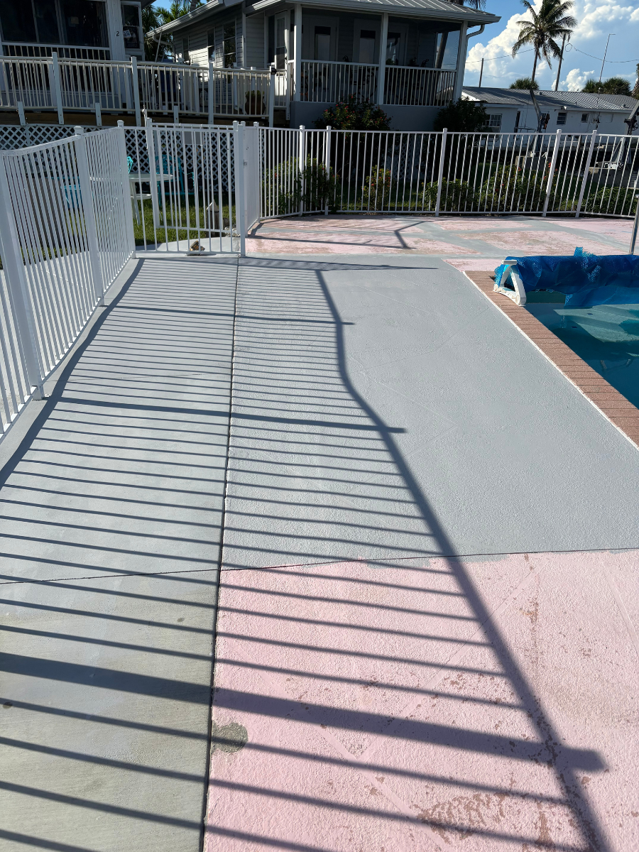 A poolside patio with a textured gray coating next to a blue swimming pool, viewed through a white metal fence.