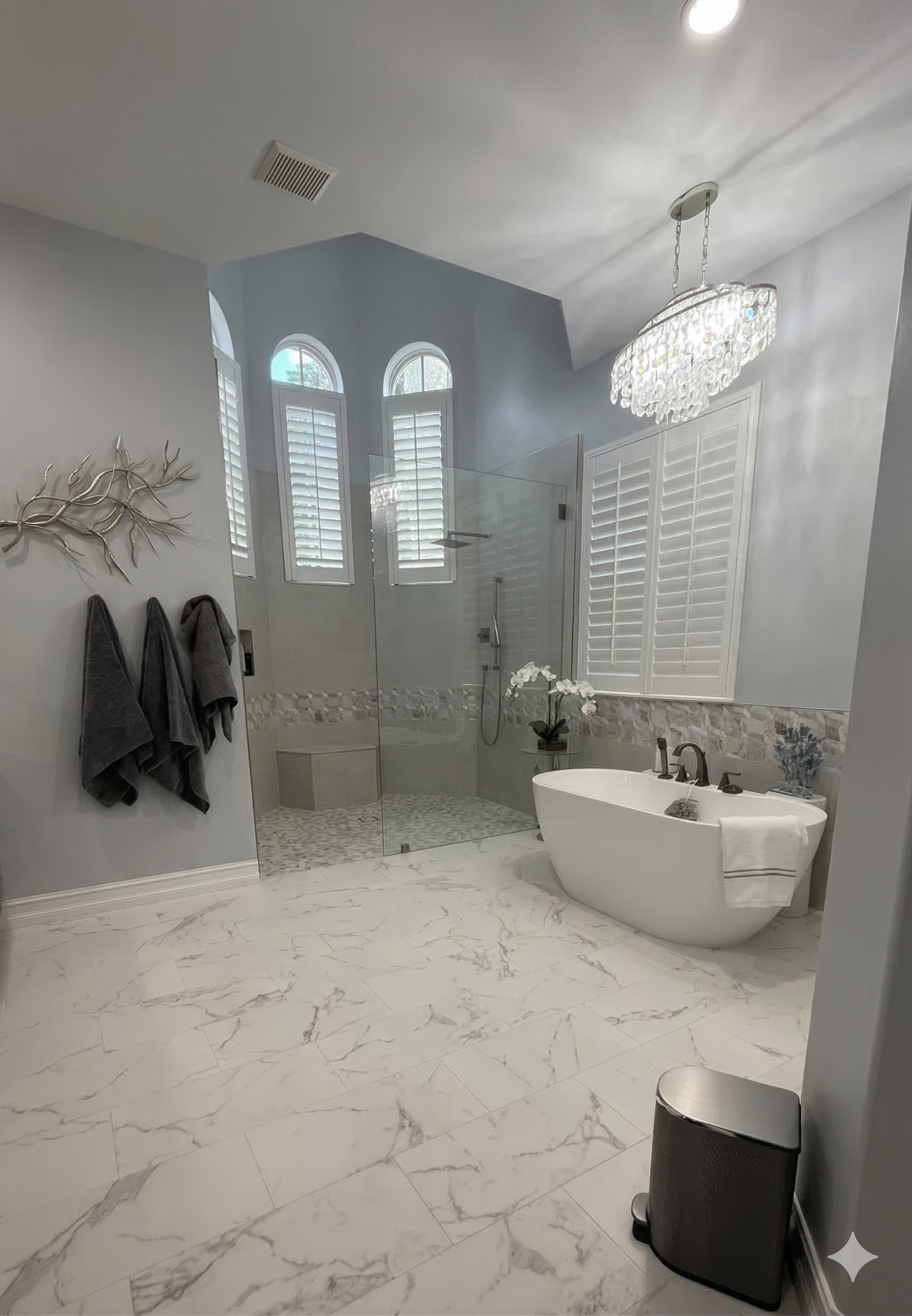 A luxurious bathroom with a white freestanding tub, a glass shower stall, marble-style flooring, and a crystal chandelier.