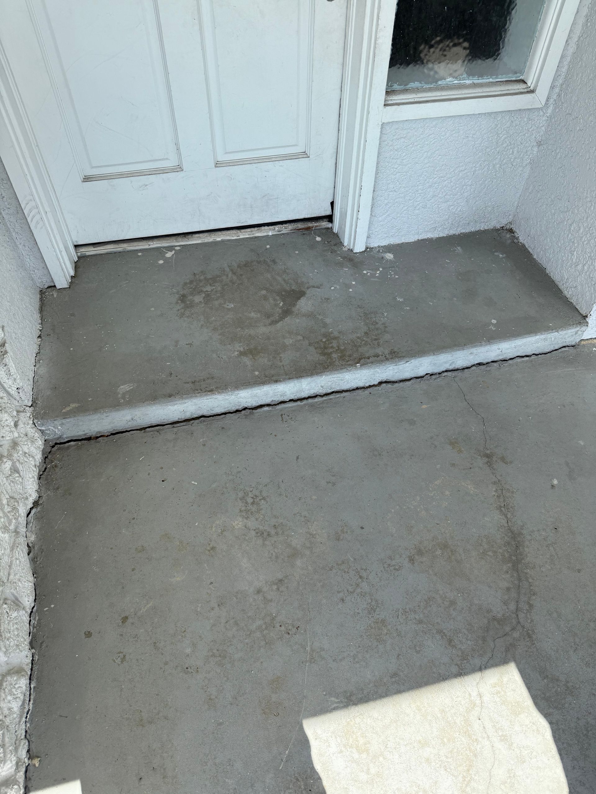 A grey concrete door step leading to a white door, with a small portion of a light-colored outdoor mat visible below.