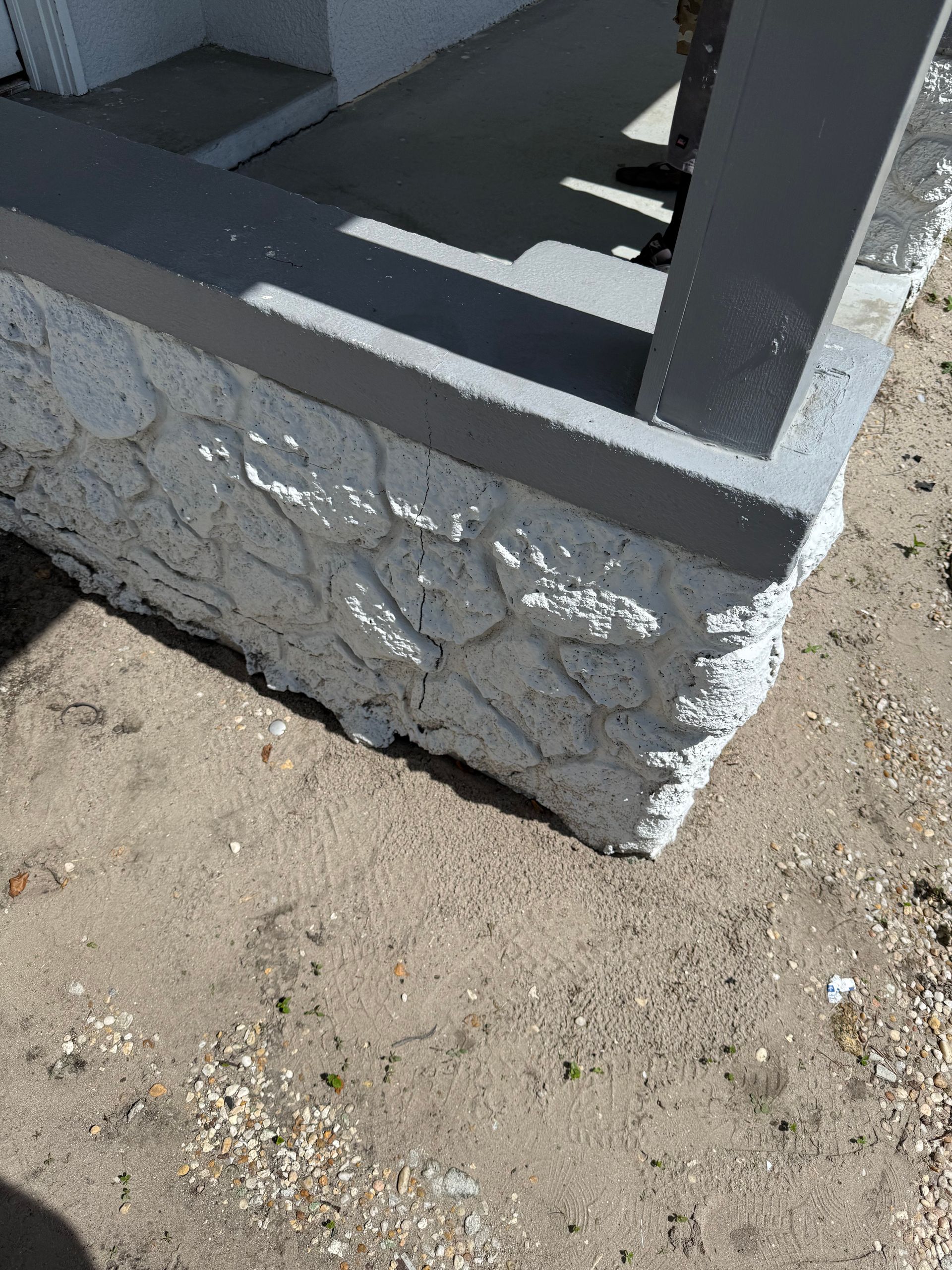 A gray porch post rises from a textured, white-painted masonry base at the edge of a sandy, dirt ground surface.