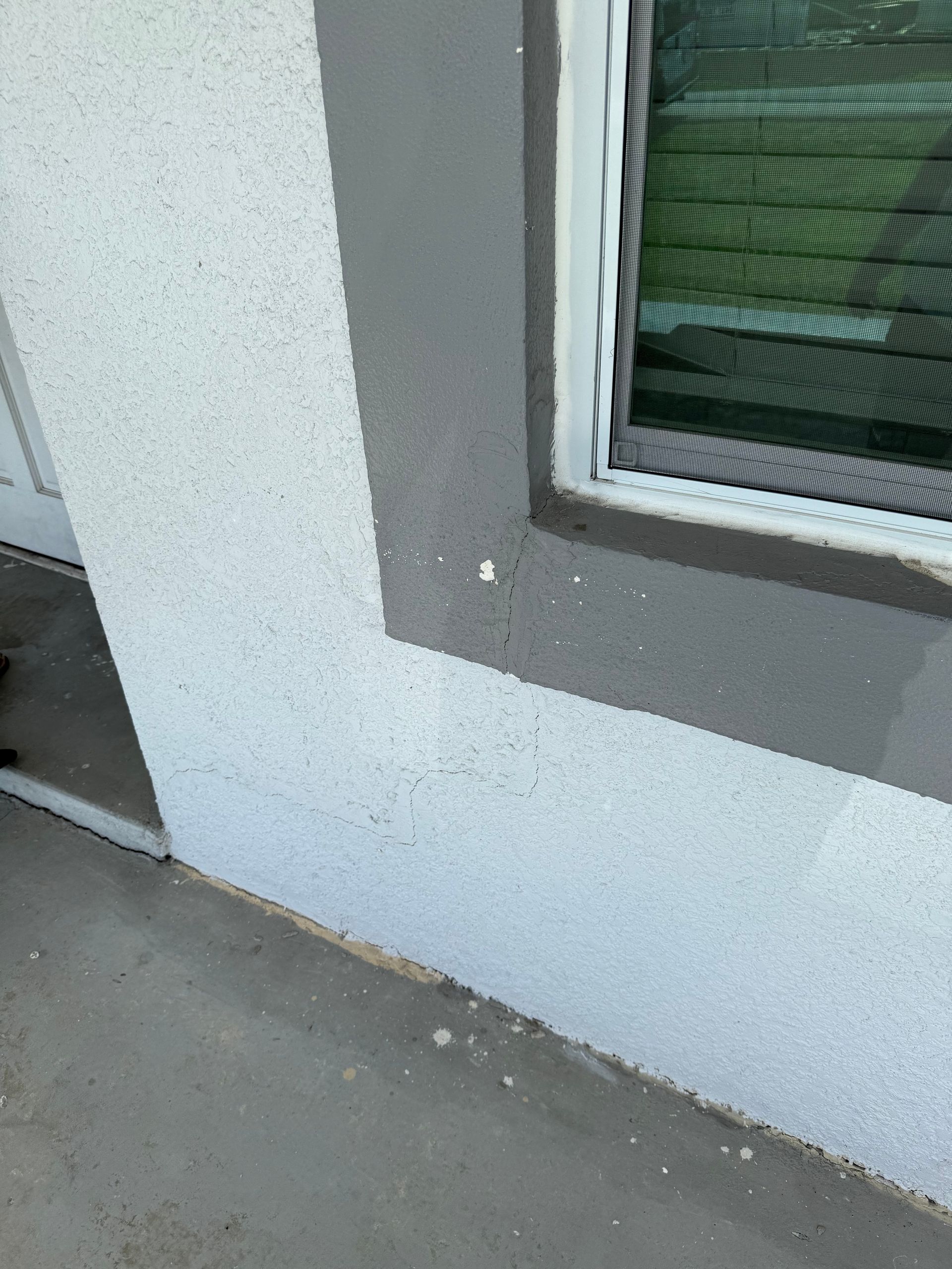 A light-colored stucco wall with a gray trim around a window next to a concrete patio.