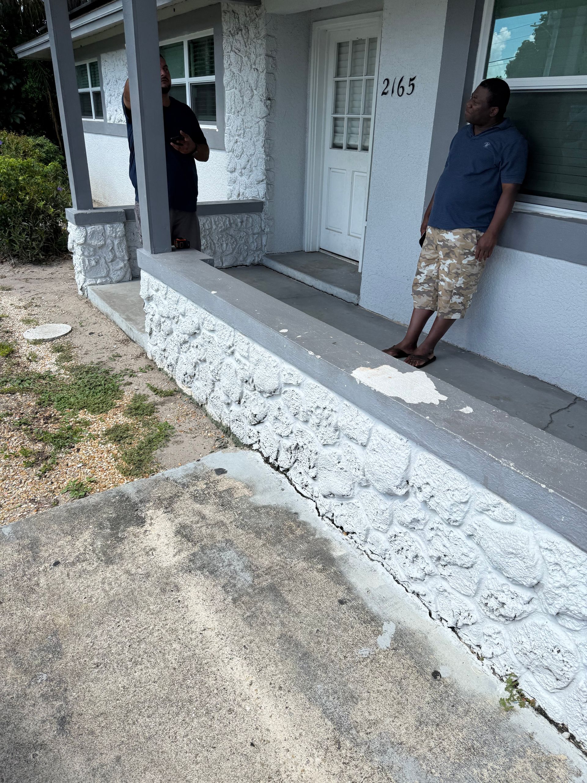 Two individuals stand on the porch of a gray house with a stone-textured base and a white front door.