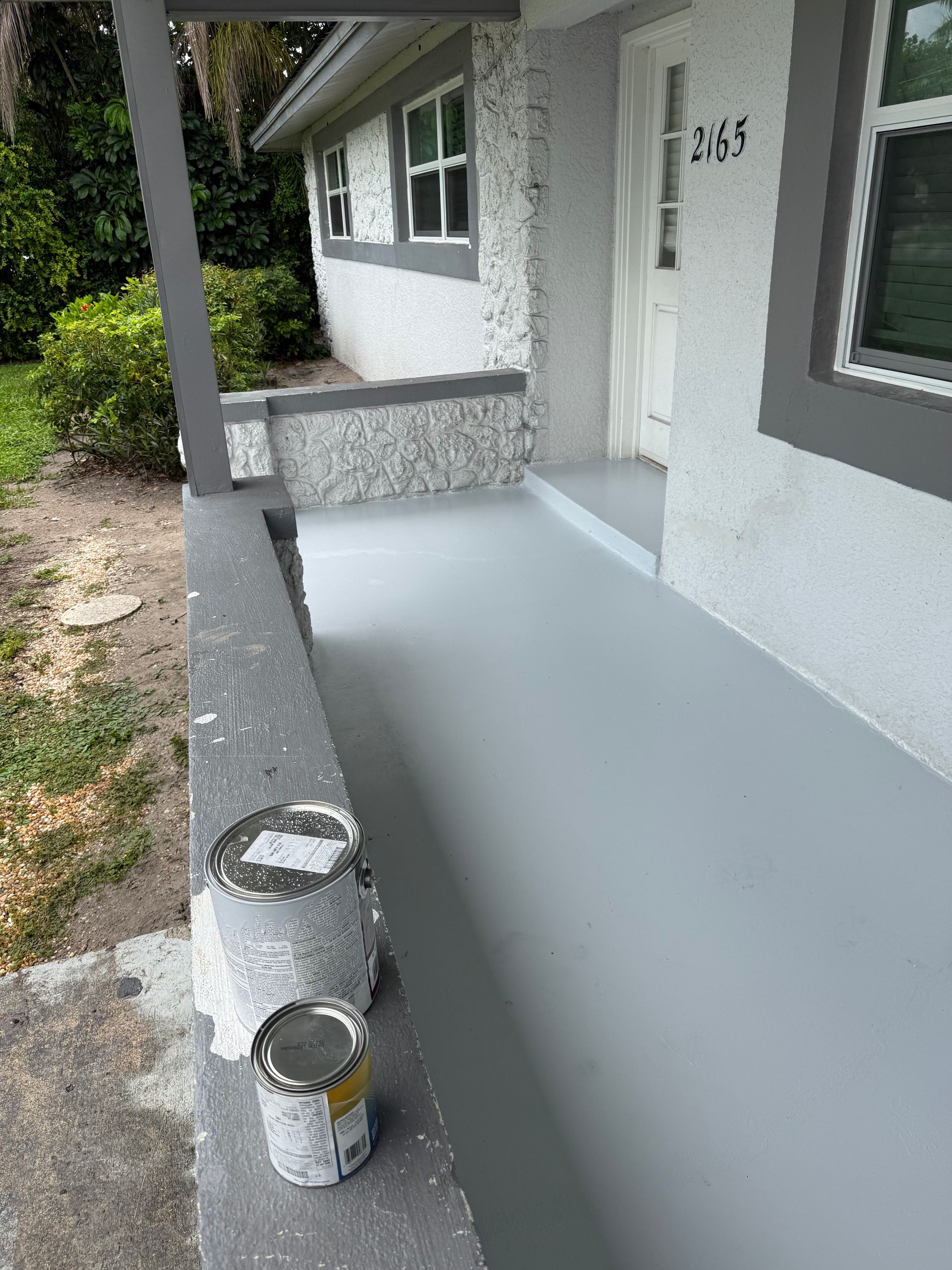 A freshly painted gray porch with two paint cans resting on the low concrete wall in front of a white house.