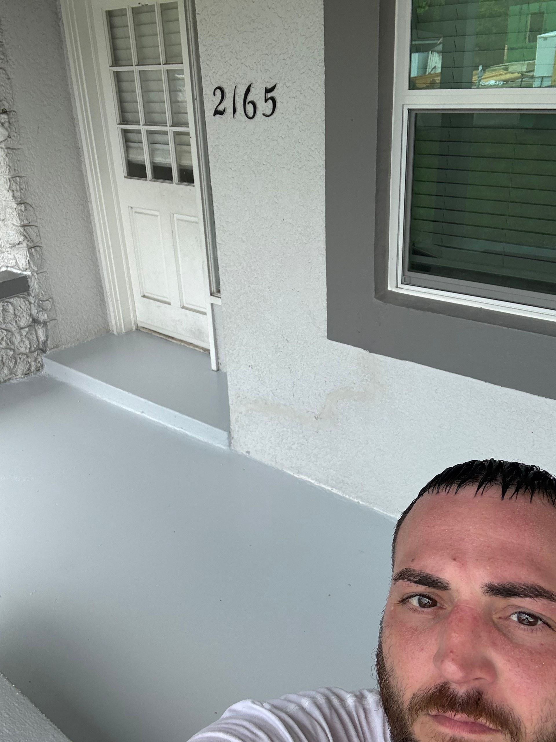 A man with dark hair and a beard takes a selfie on a porch with a white door labeled 2165 and gray painted concrete.