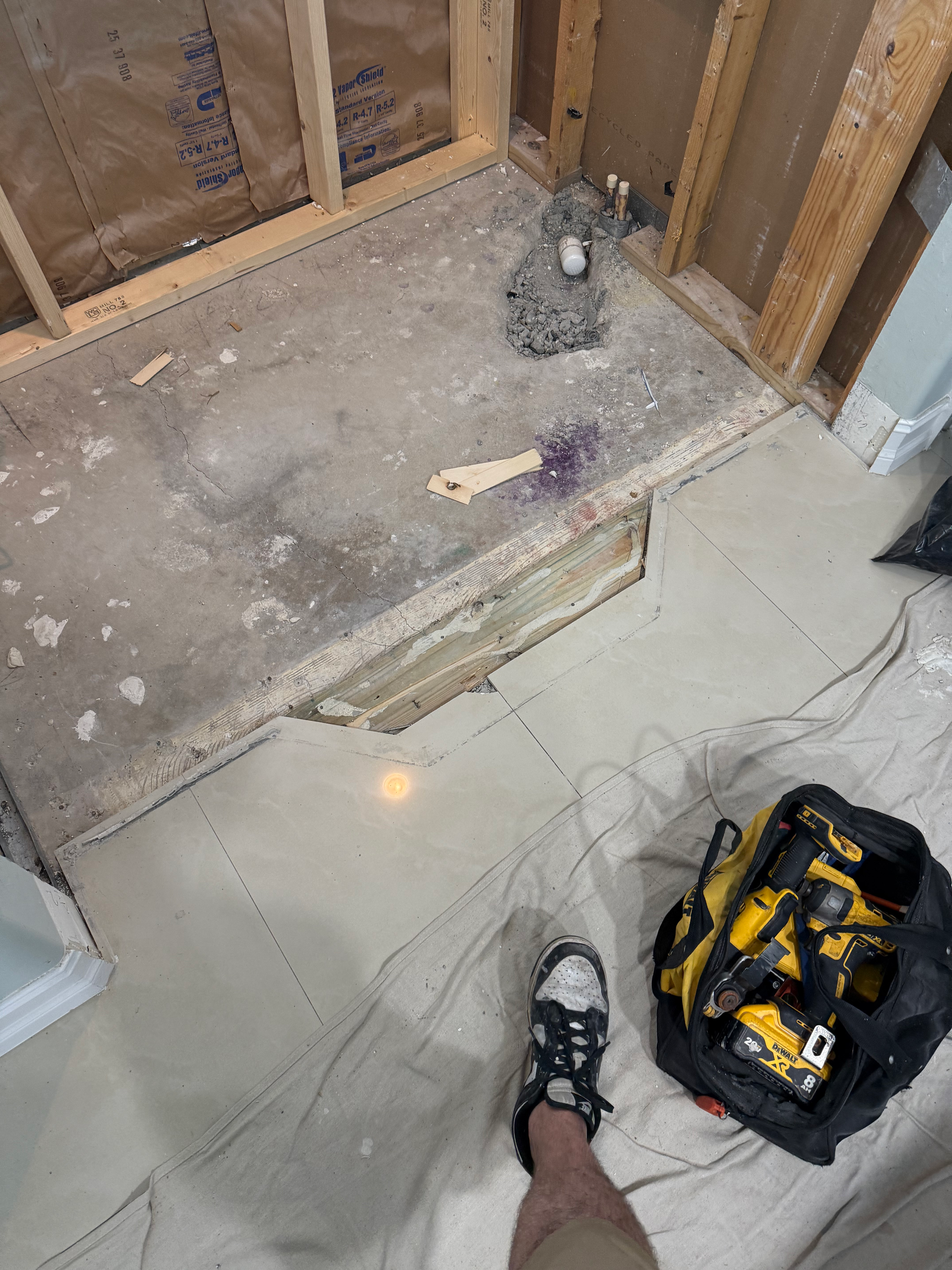 An interior construction site showing a shower base framed with wood studs, concrete flooring, and a tool bag on the floor.