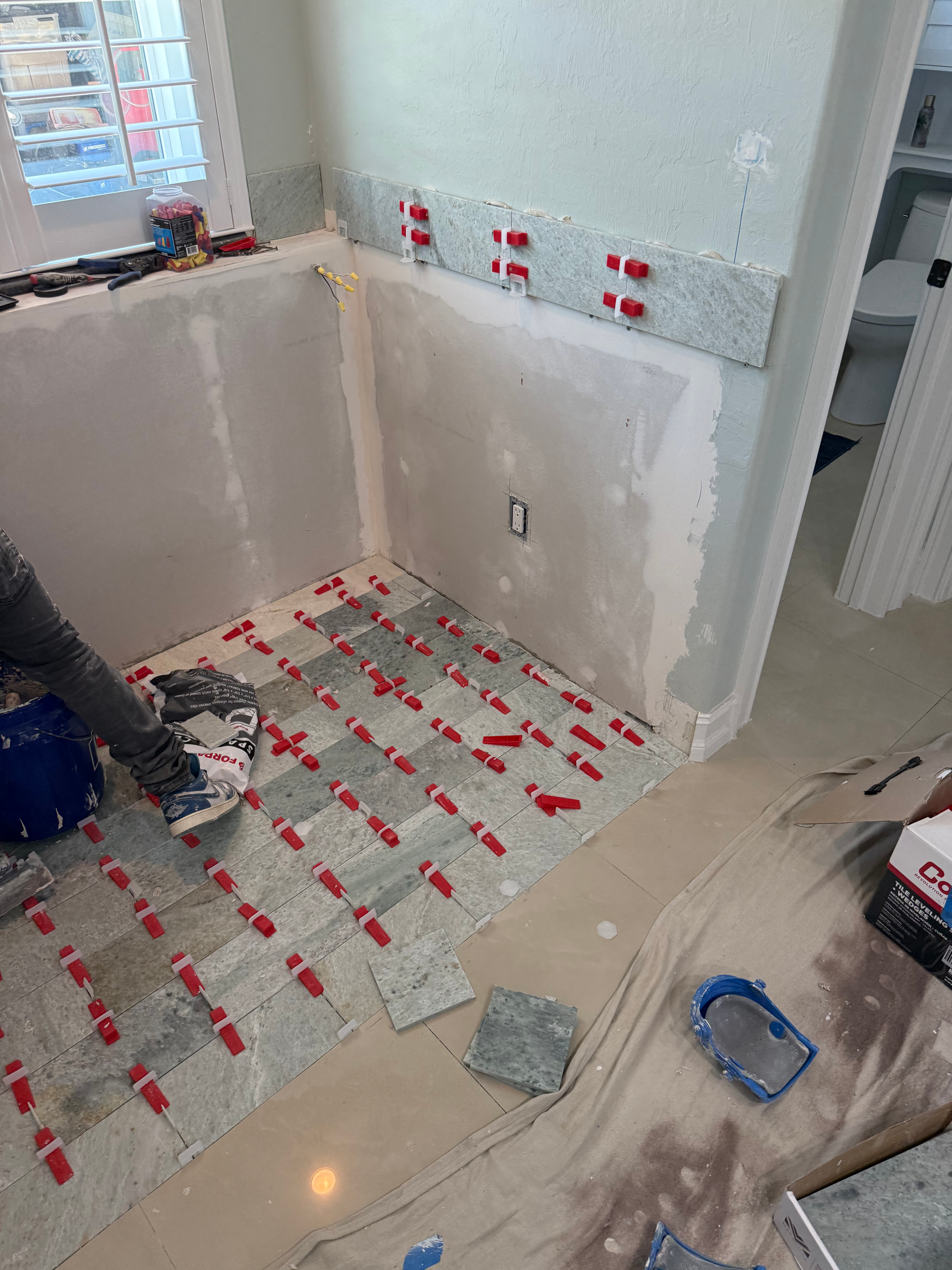 A person installing stone tiles on a floor and wall in a bathroom, using red plastic leveling spacers.