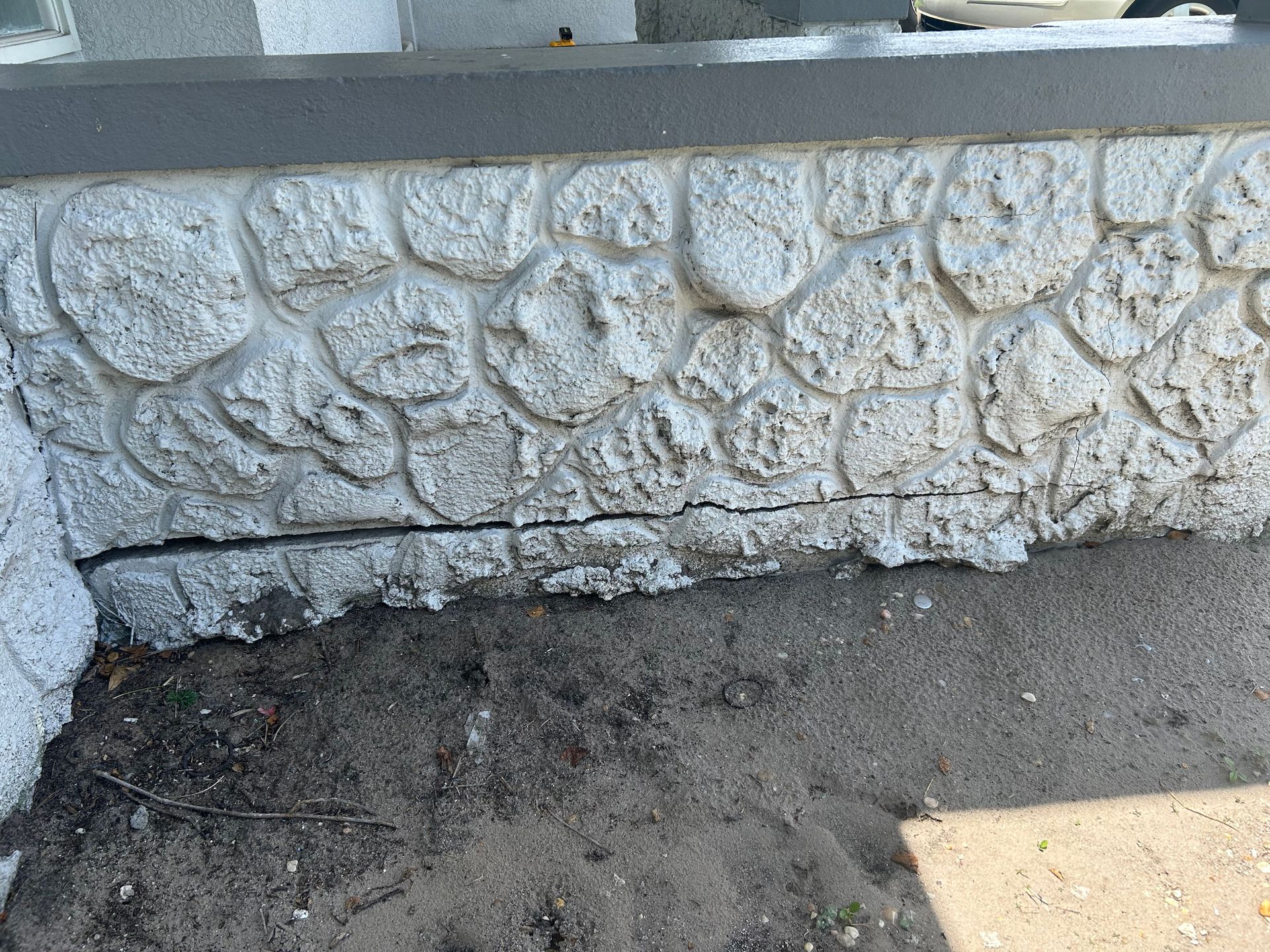 A horizontal crack runs along the base of a white, textured stone-patterned concrete wall above dirt and pavement.
