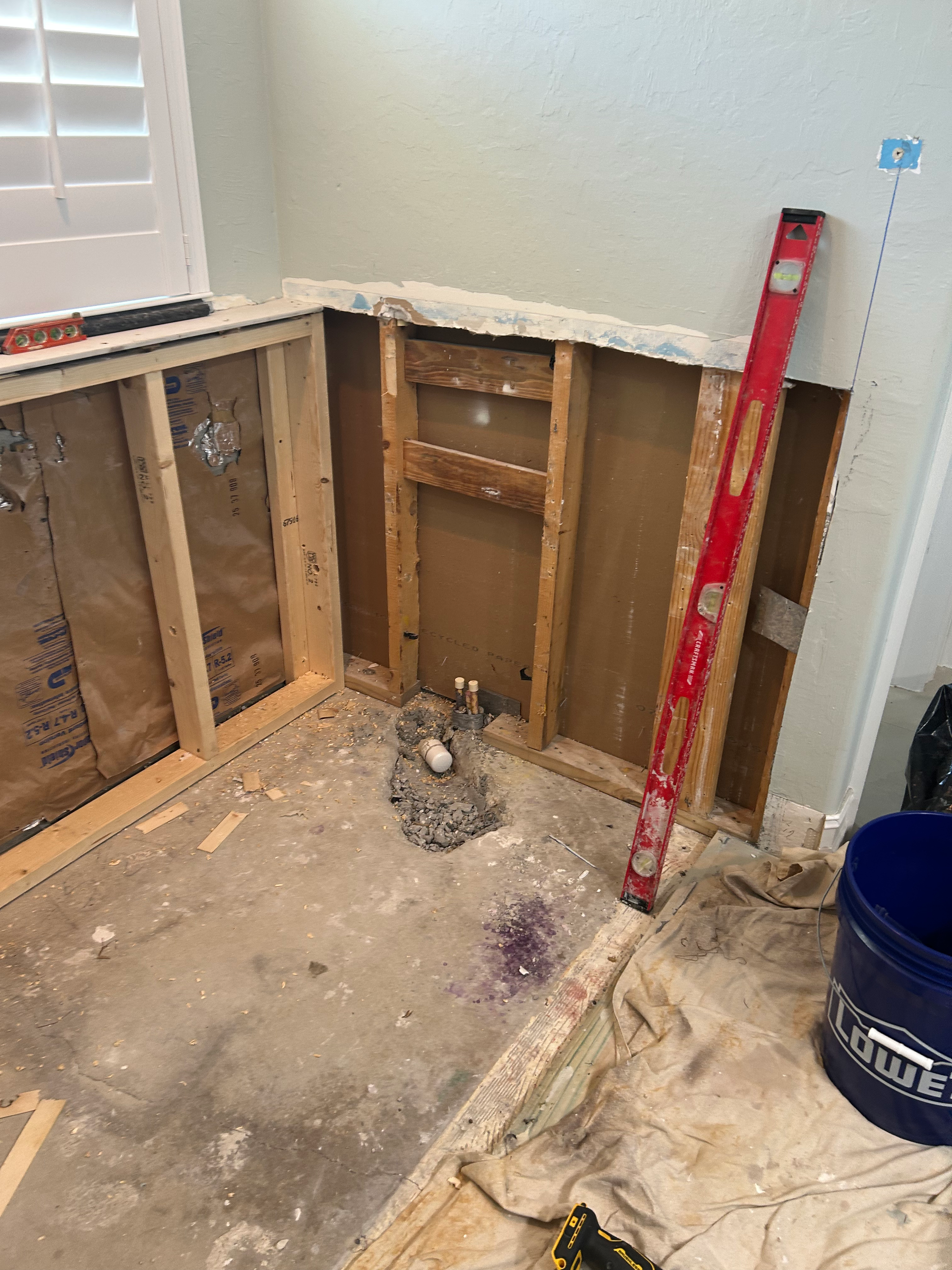 Construction area in a home showing exposed wooden wall studs, insulation, a plumbing drain, and a red level tool.