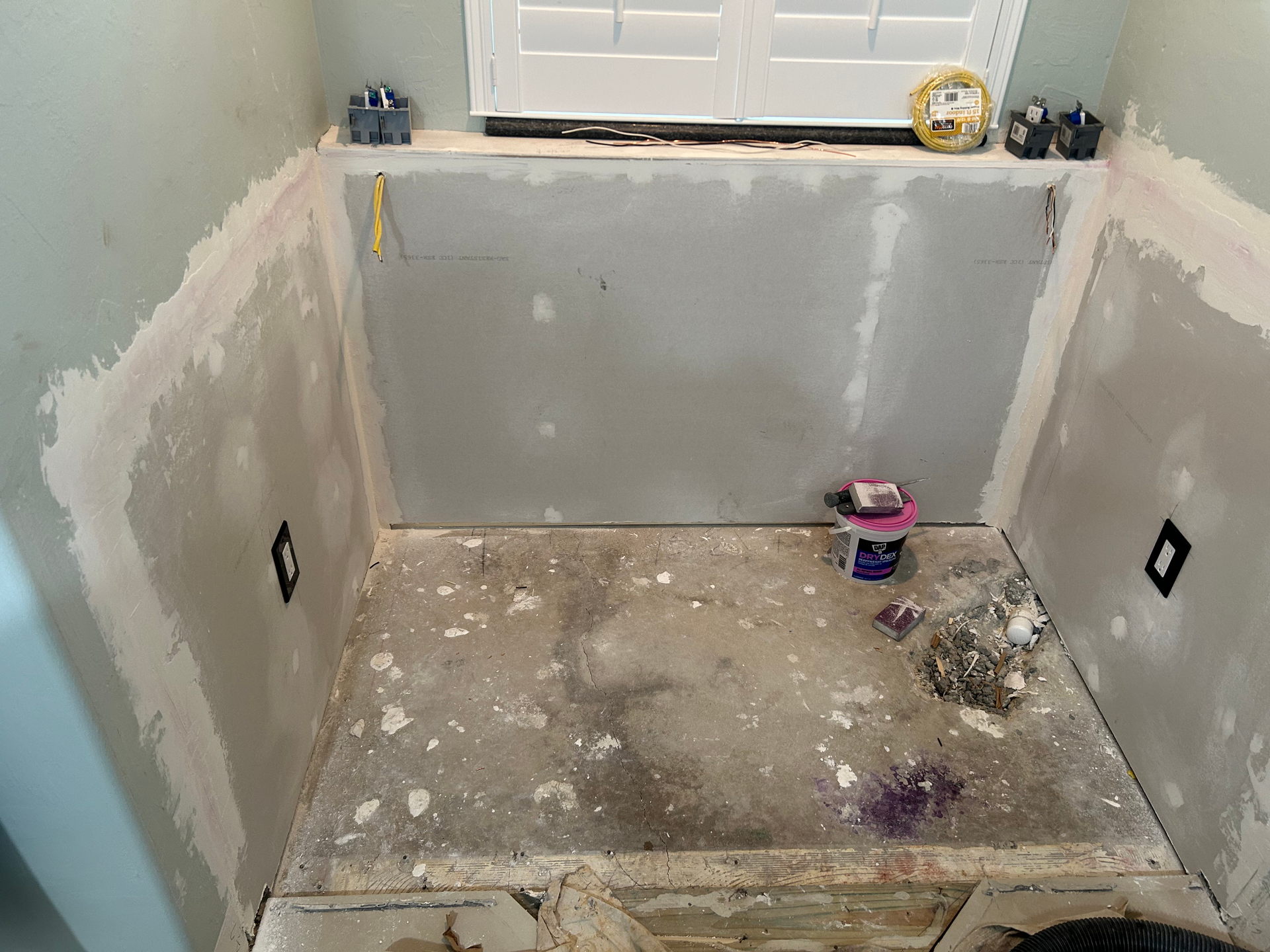 Drywall installation in an alcove with taped seams, two electrical boxes, and debris on the floor beneath a window.