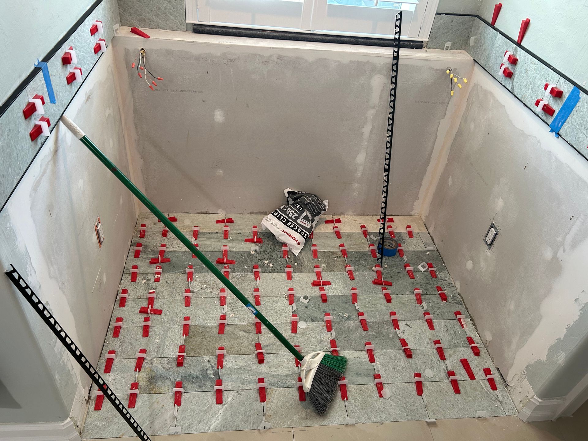 A bathroom floor under renovation with gray stone tiles, red leveling spacers, and a broom leaning against the wall.