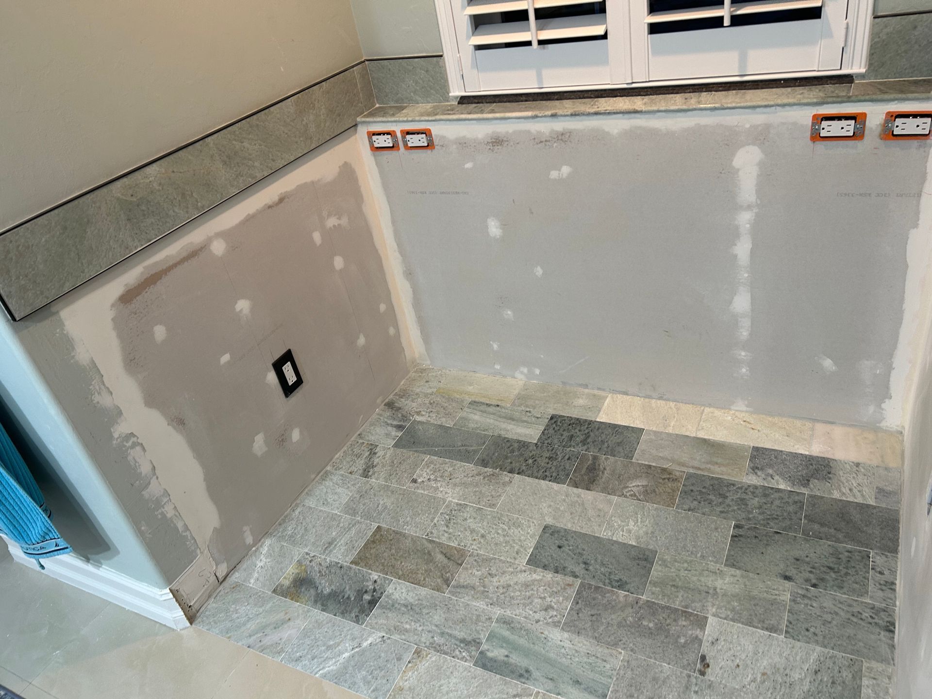 A room corner undergoing renovation with unfinished drywall, electrical outlets, and grey stone floor tiling.