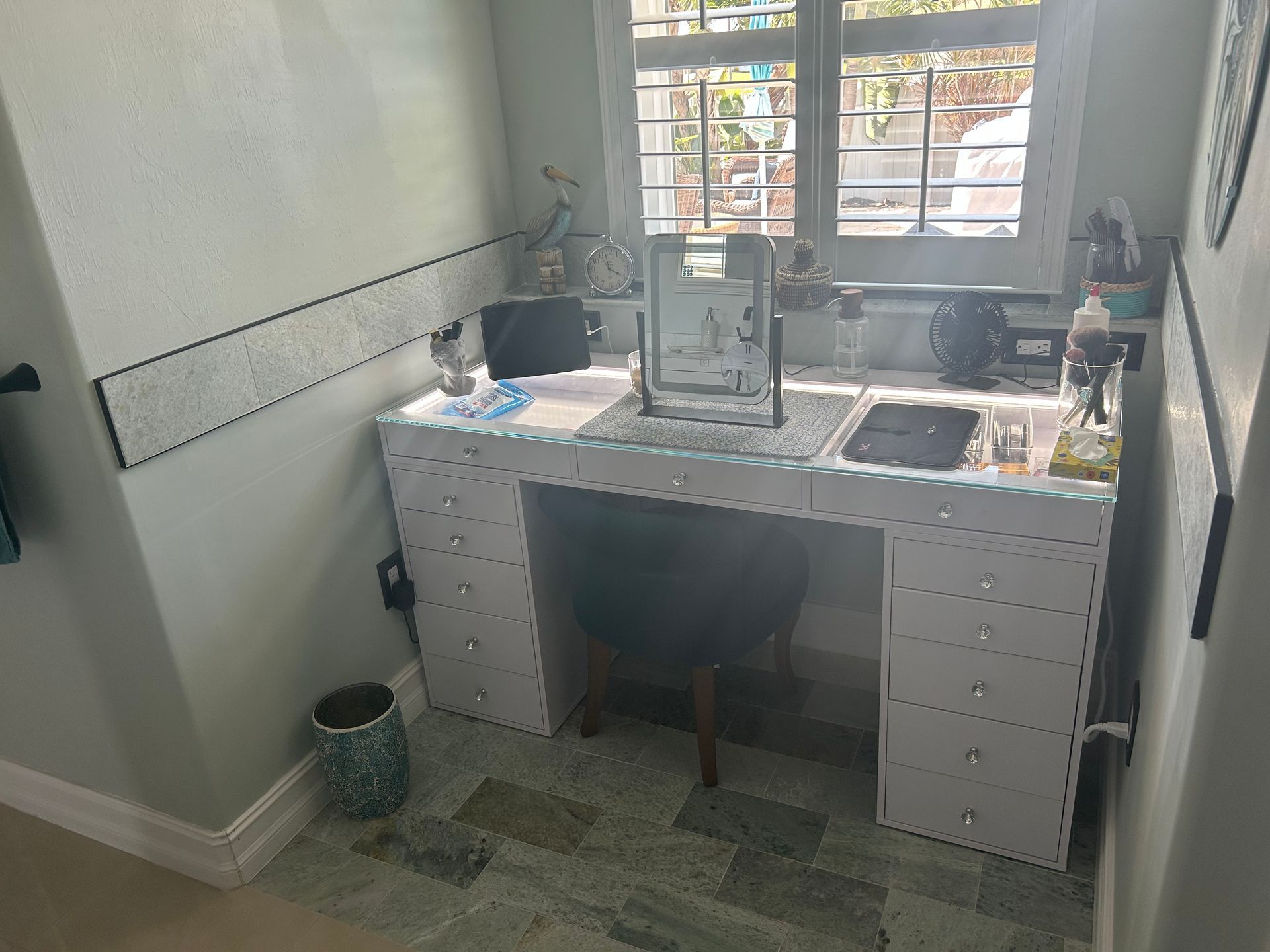 A white vanity desk with multiple drawers, a mirror, and a small chair sits under a window in a bright room.