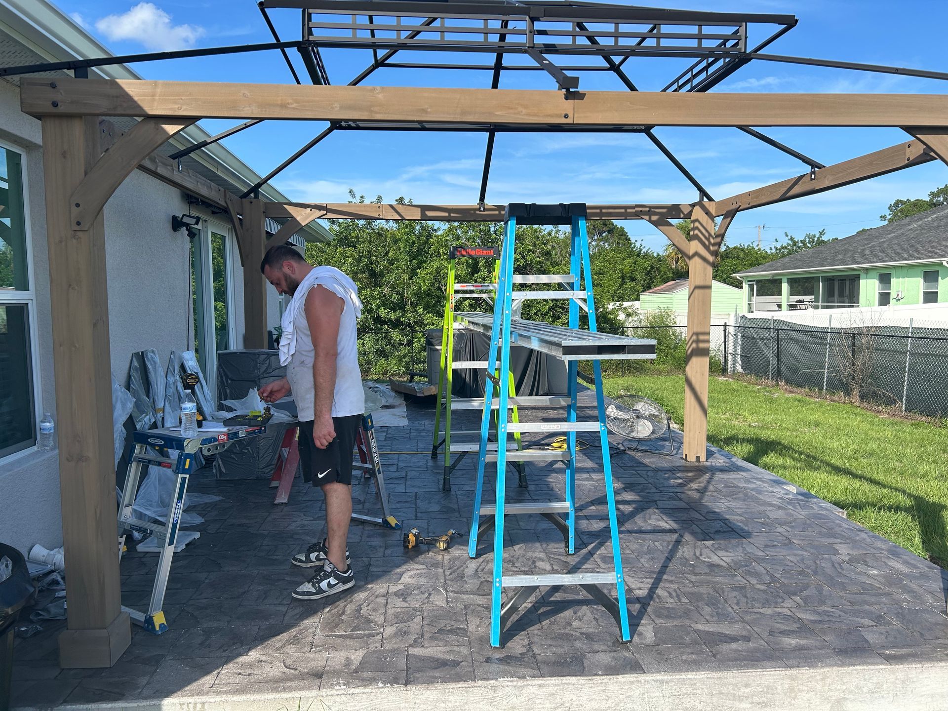 A person working on an outdoor patio under a wooden gazebo frame, with ladders and tools nearby on a sunny day.