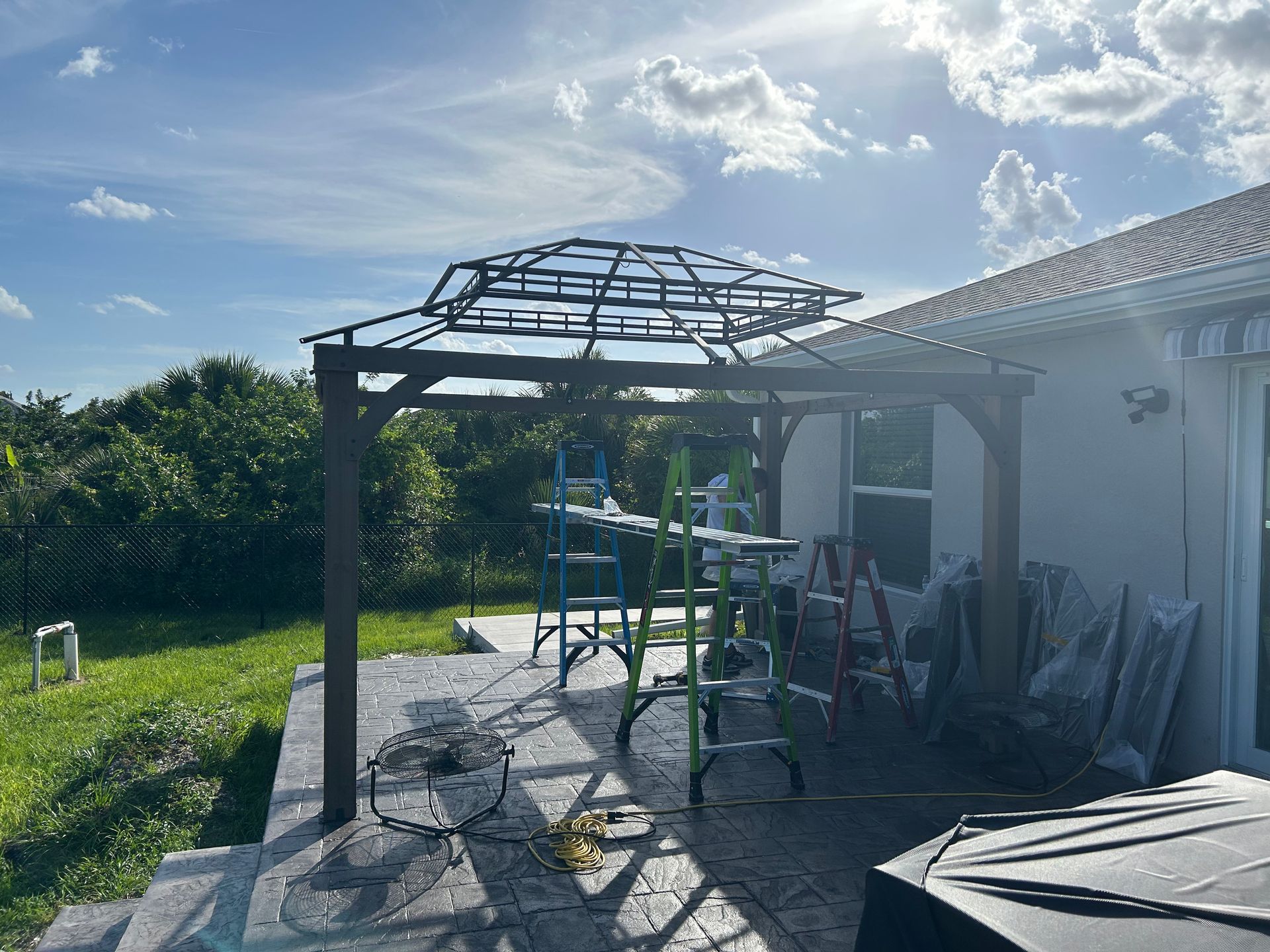 A wooden gazebo frame under construction on a sunny backyard patio with step ladders and equipment nearby.