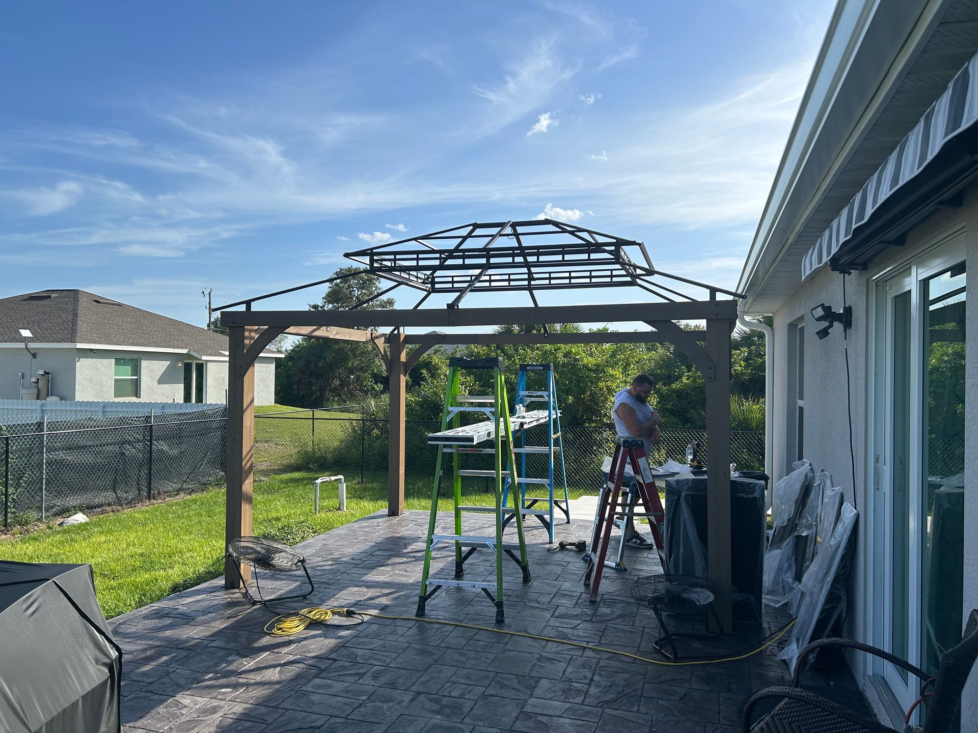 A person stands on a ladder assembling a metal gazebo frame on a residential patio under a bright, clear blue sky.