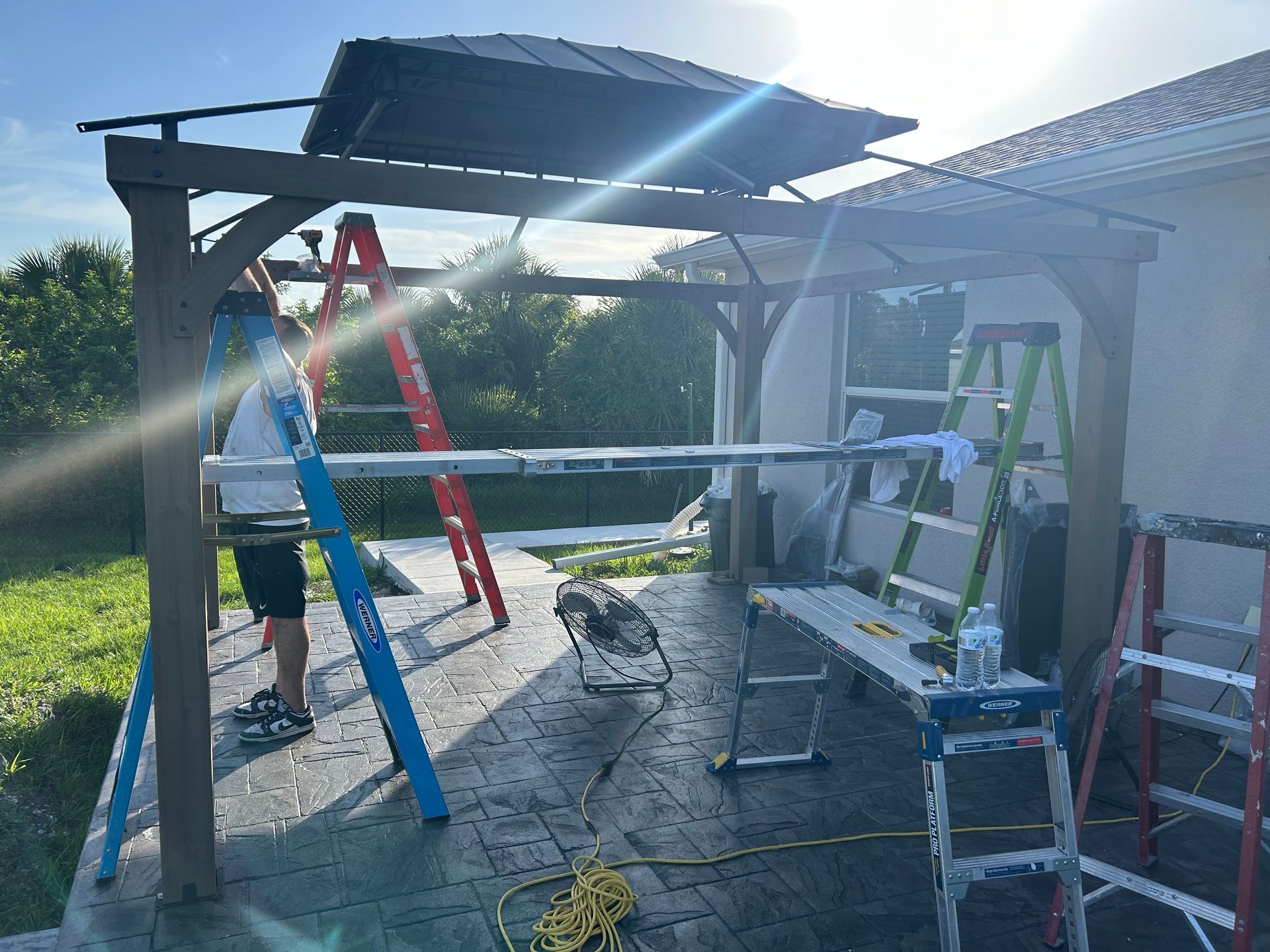 A person in work clothes stands on a blue ladder under a backyard gazebo frame with multiple ladders and tools.