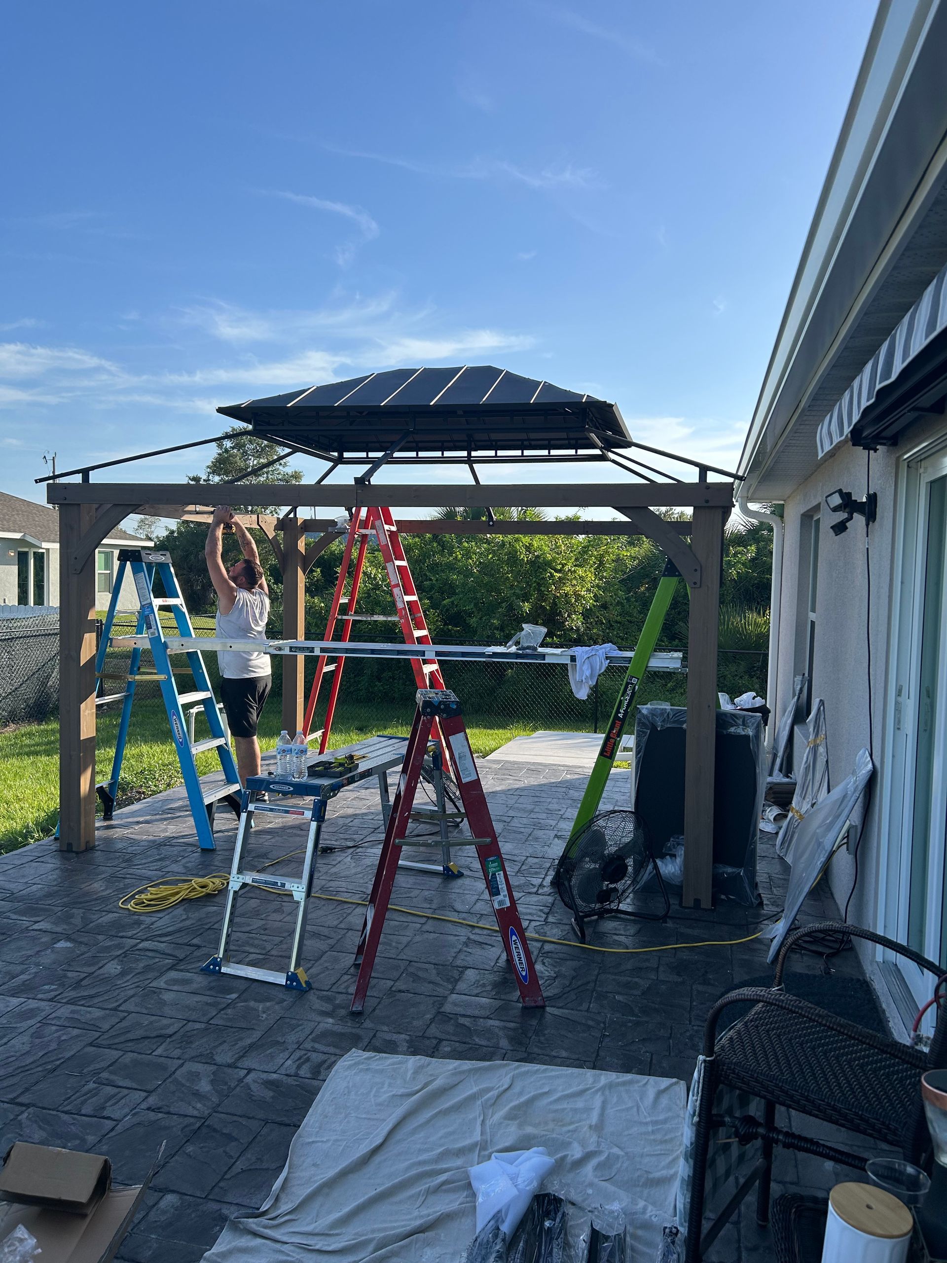 A person stands on a blue ladder while assembling a wooden gazebo frame on a stone patio under a bright, clear blue sky.