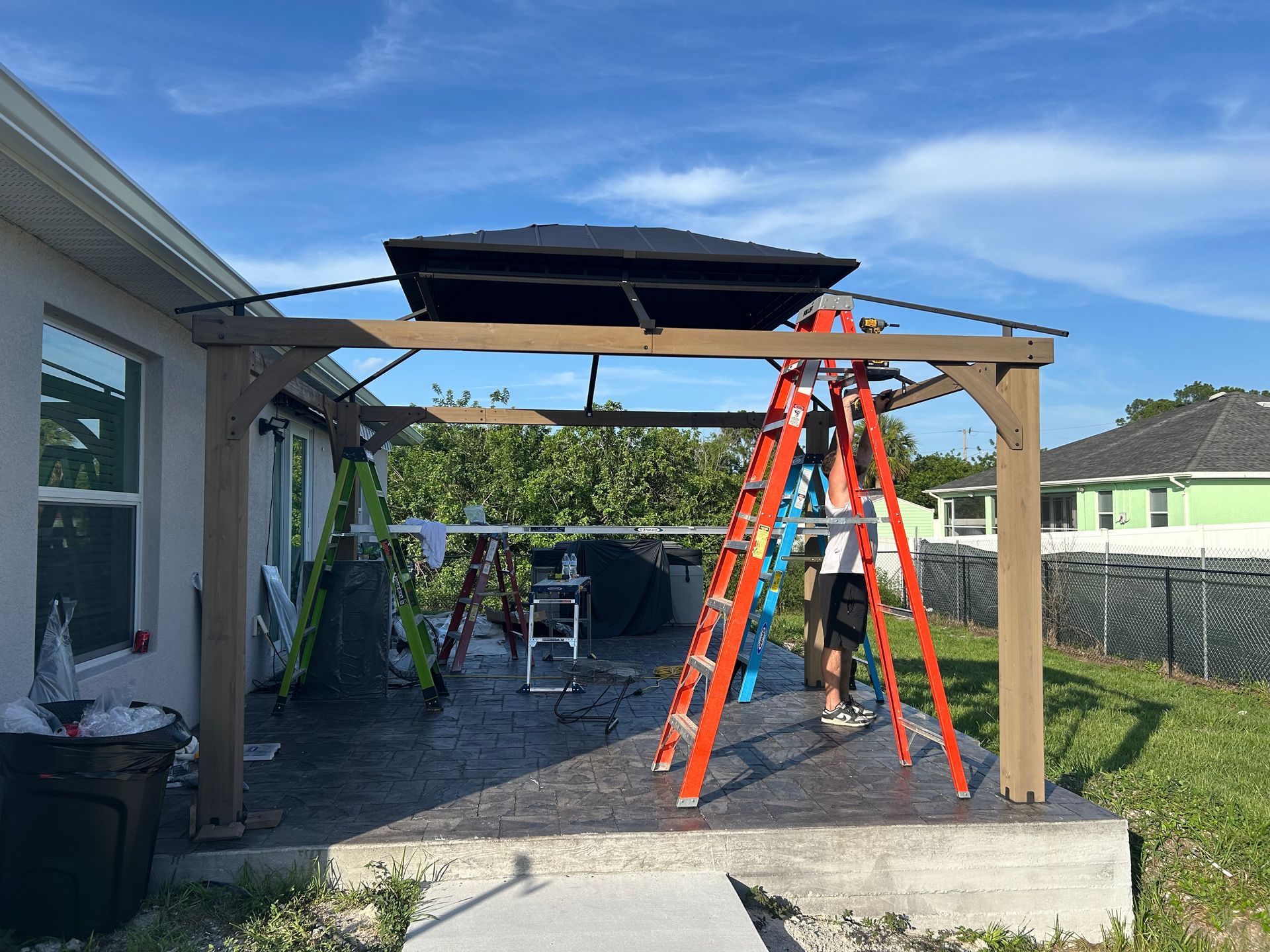 A person on a ladder installs a gazebo frame on a residential patio under a bright blue sky.
