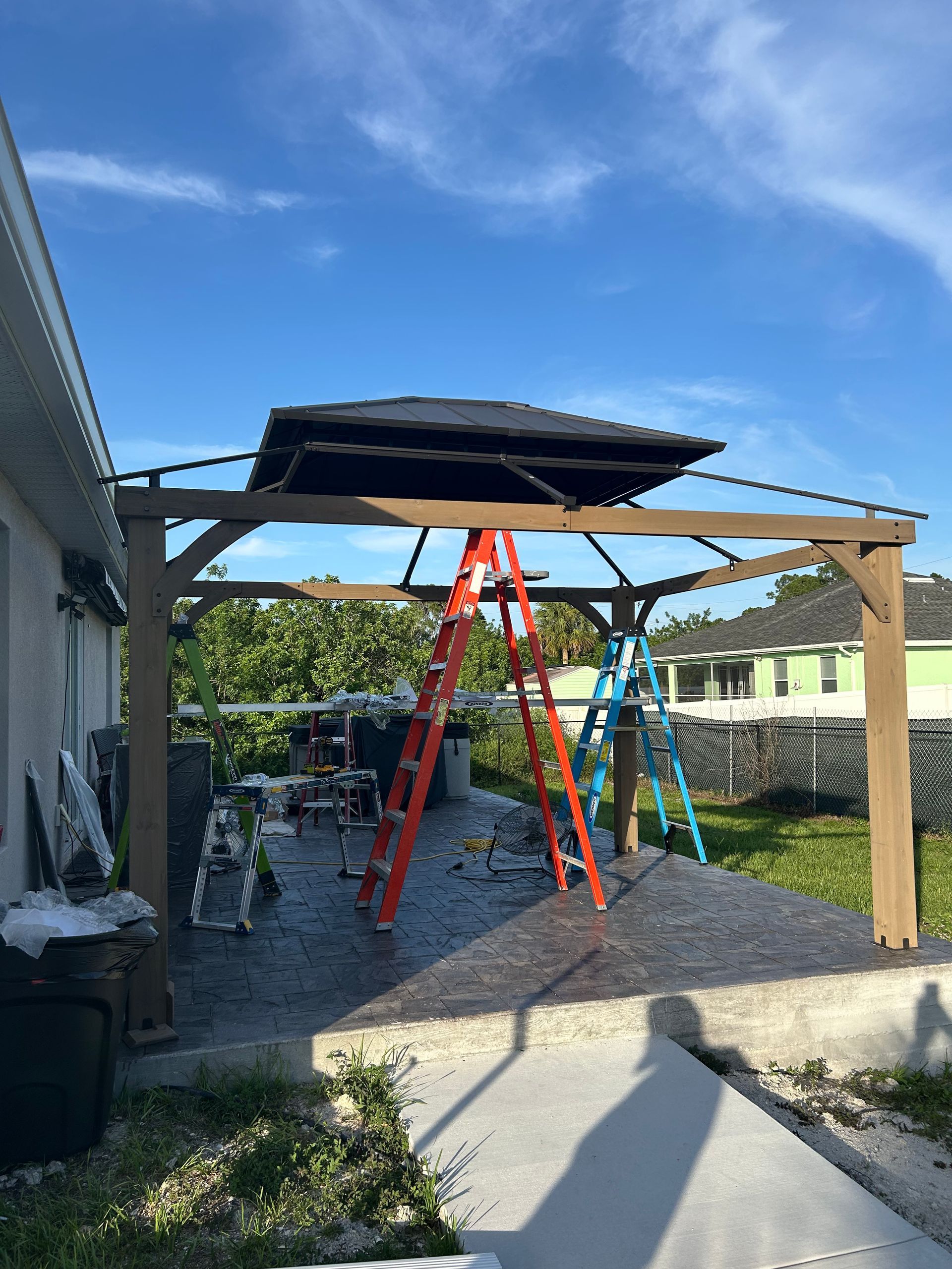 A partially assembled outdoor gazebo with a black roof sits on a patio, supported by wooden beams, with ladders nearby.