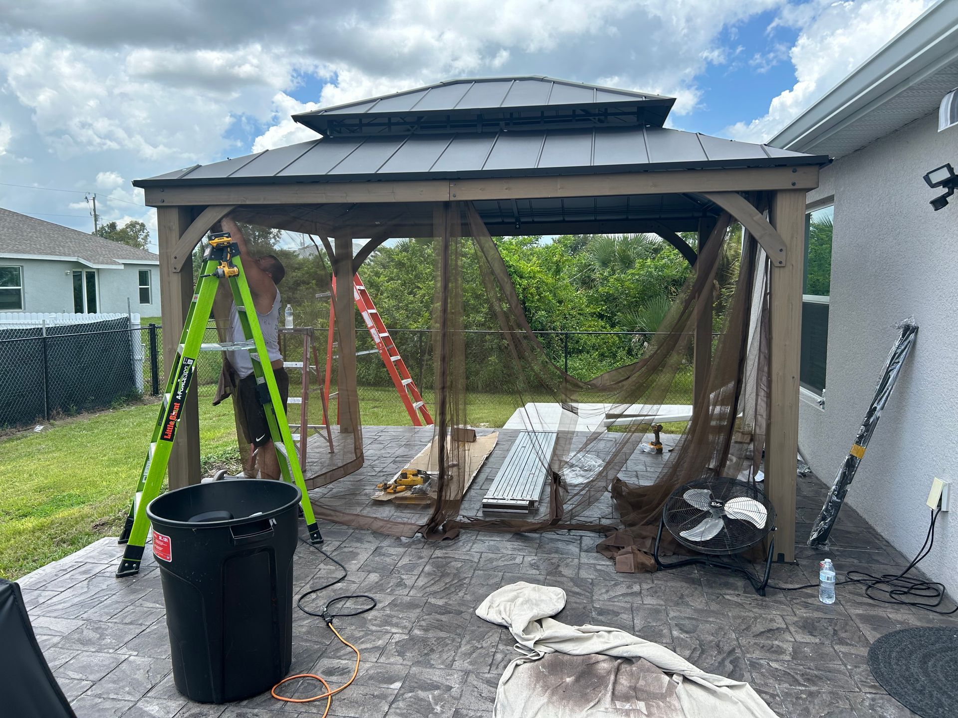 A person stands on a green ladder painting a gazebo frame on a residential patio.