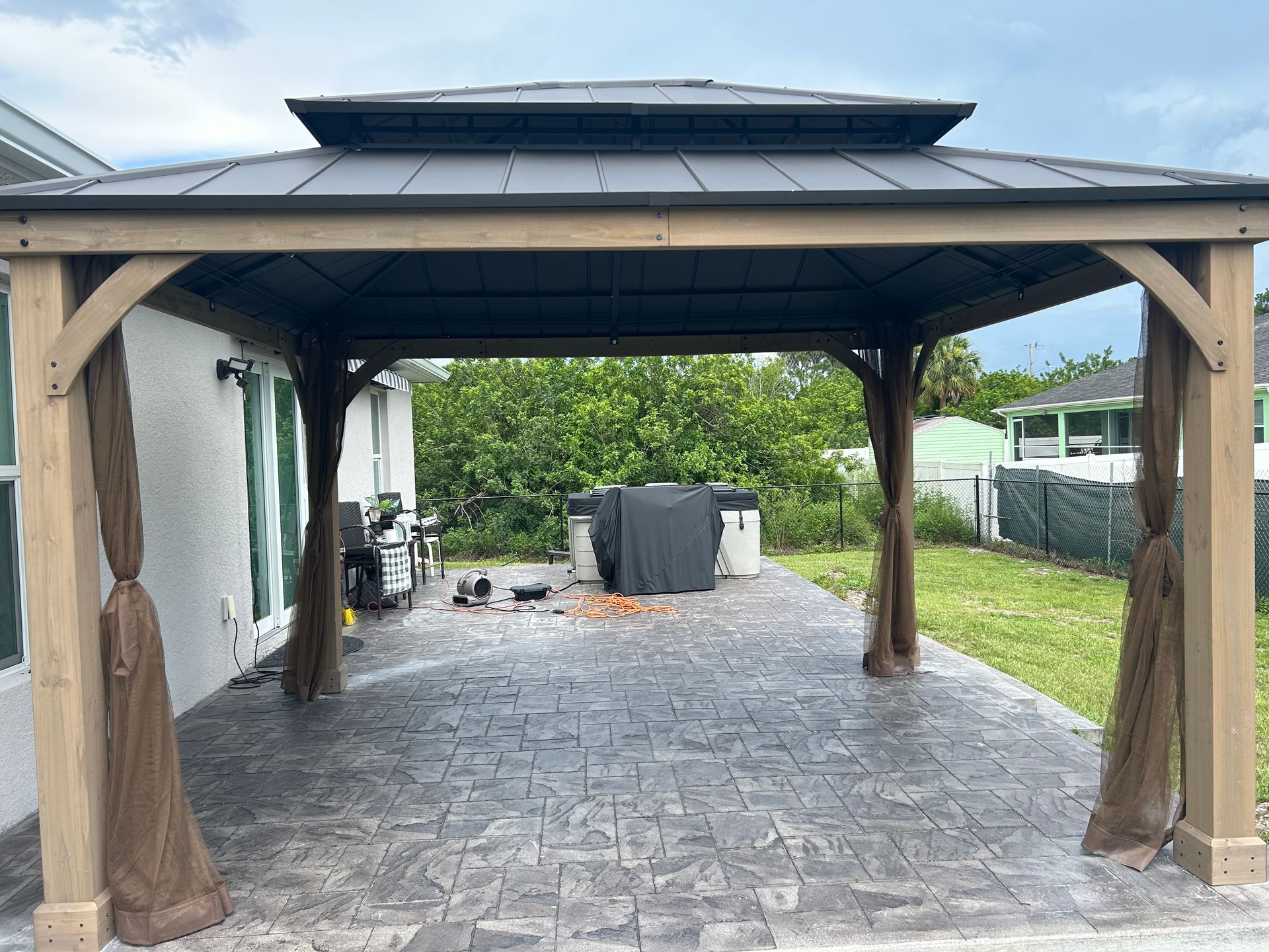 A wooden-frame gazebo with a dark metal roof, set on a gray stone patio in a backyard.
