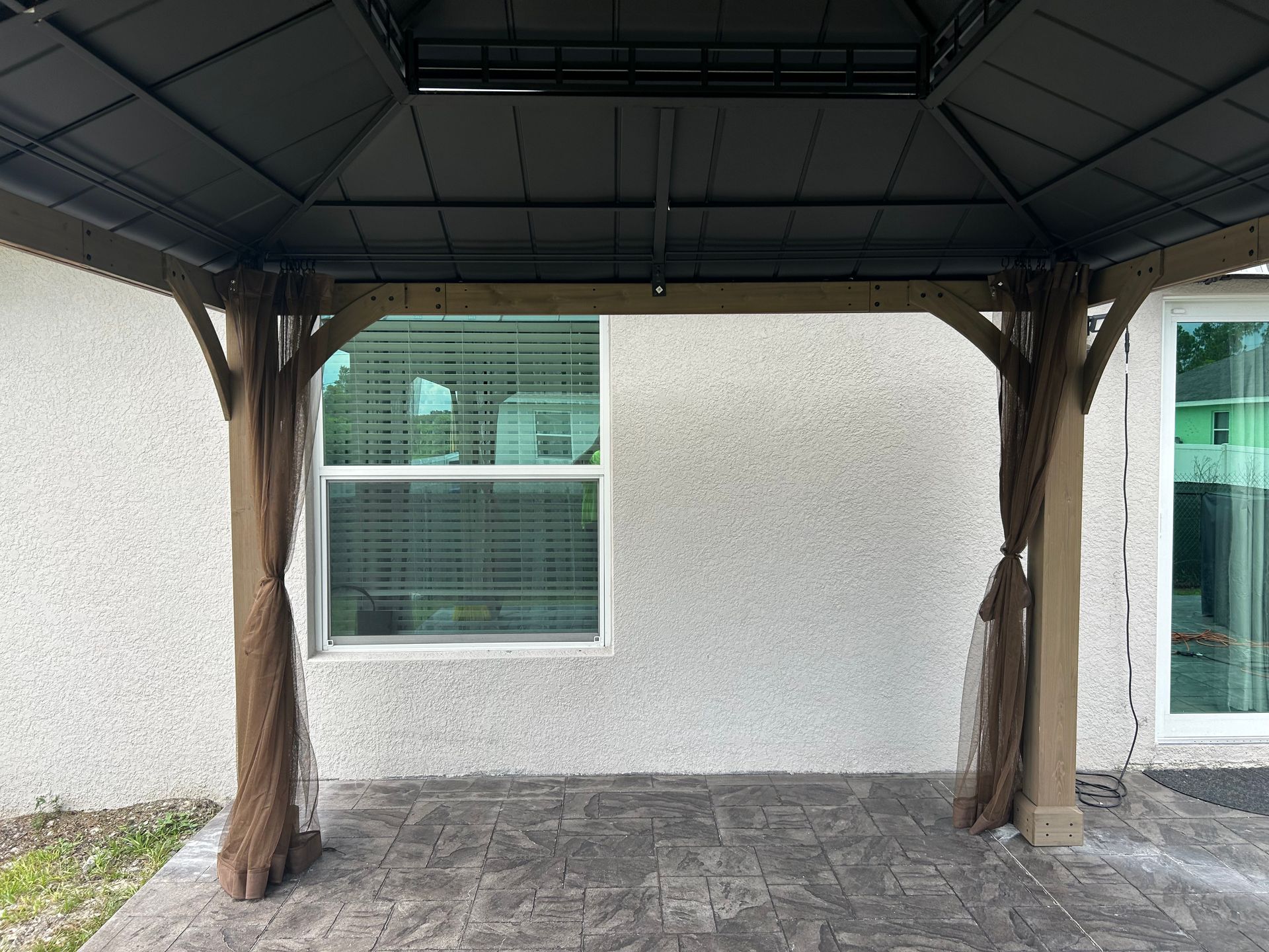 A patio gazebo with a black metal roof and brown curtains, positioned against a light-colored stucco house wall.