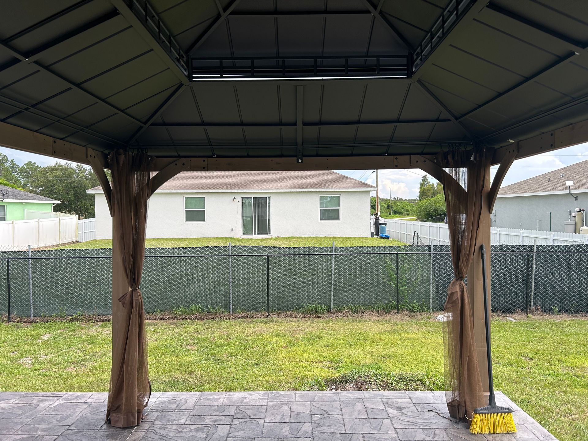 A dark brown metal gazebo with sheer curtains stands on a stone-patterned patio, overlooking a grassy yard and white house.