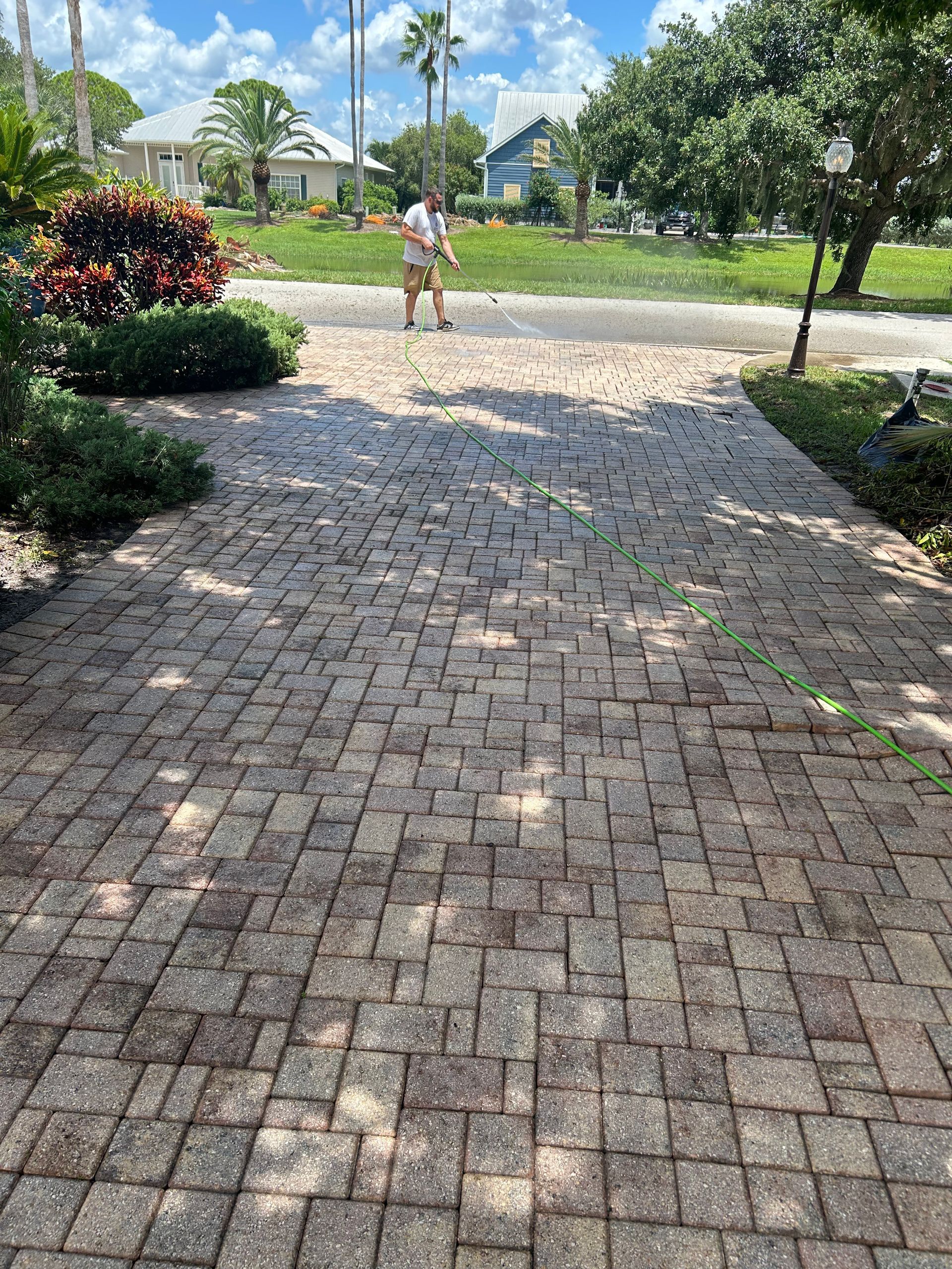 A person uses a leaf blower to clean a brick driveway on a sunny day with houses and palm trees in the background.